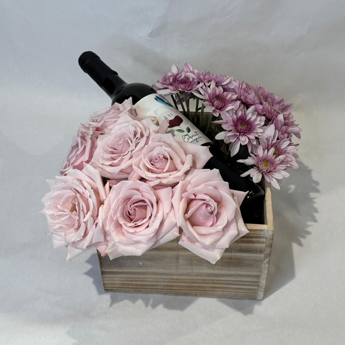 A floral gift box featuring blush roses and lavender mums arranged around a wine bottle in a wooden crate. Wine not included.