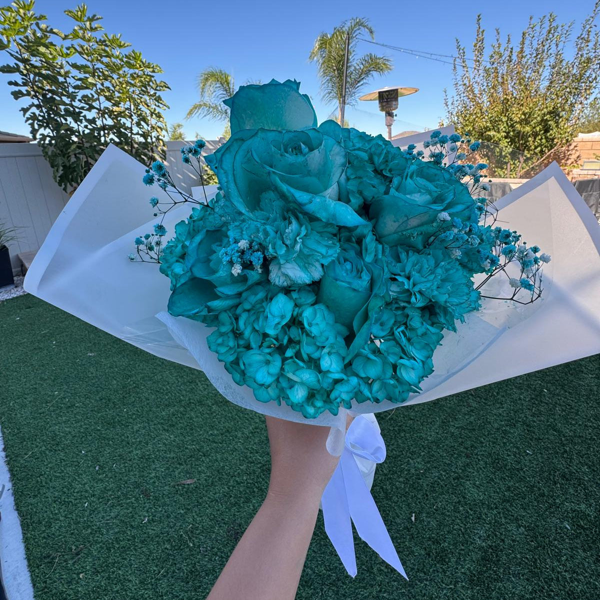 A monochromatic turquoise bouquet featuring fully turquoise-dyed blooms arranged tightly in a round, dome-style shape. The bouquet is wrapped in crisp white translucent paper and tied with a white satin ribbon, displayed on a white stand against a dark surface.