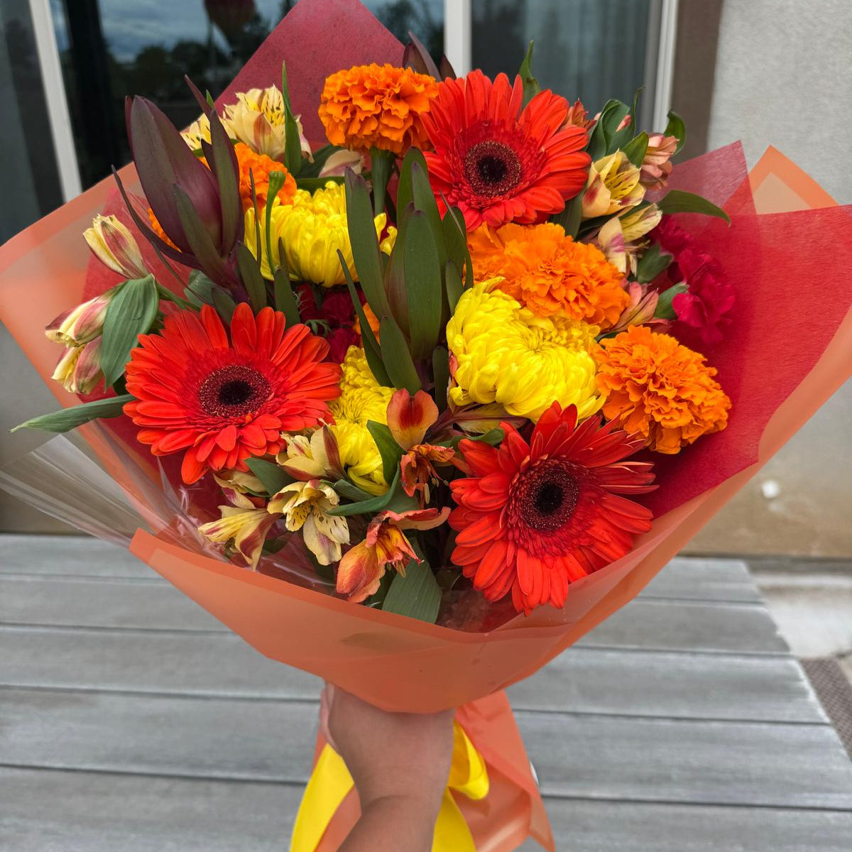 Bright fall bouquet with orange gerbera daisies, yellow mums, marigolds, and alstroemeria wrapped in autumn-colored paper with a yellow ribbon.