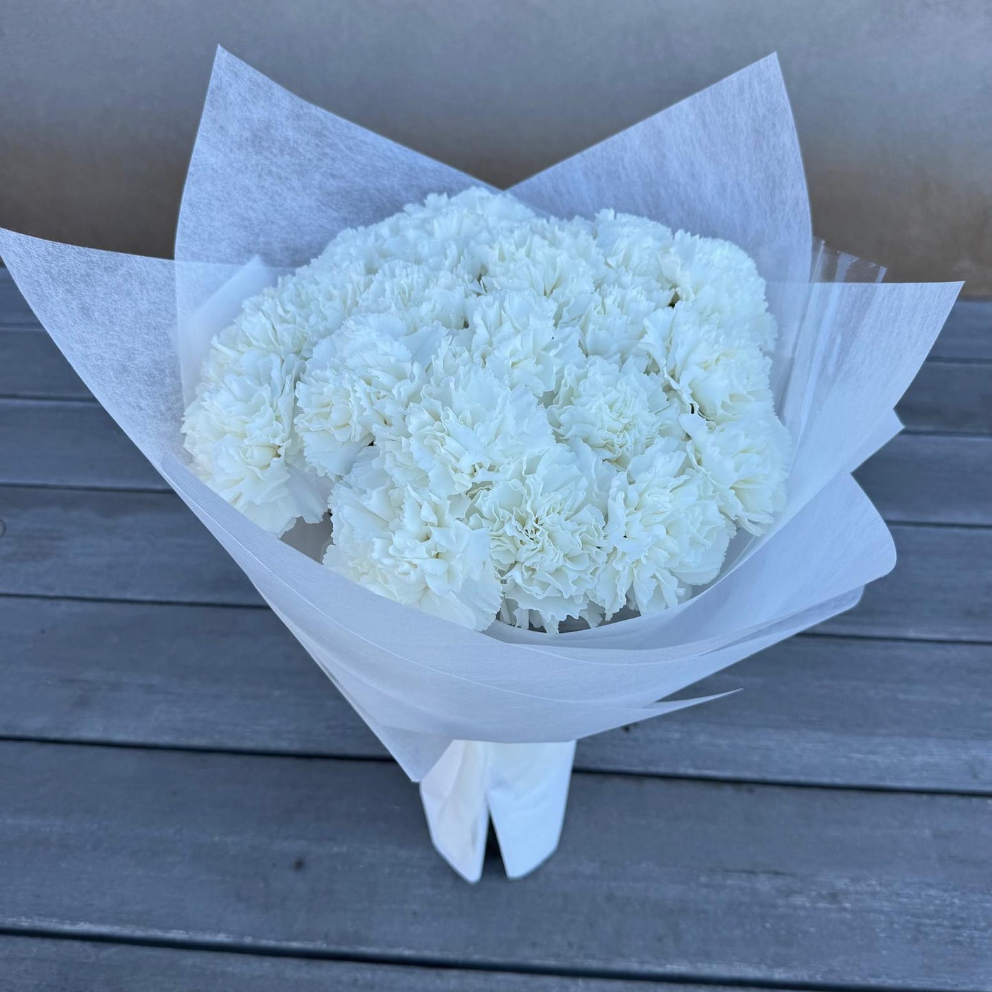 A bouquet of pure white carnations wrapped in crisp white paper, placed on a gray wooden surface. The flowers are tightly arranged, creating a soft and full appearance.