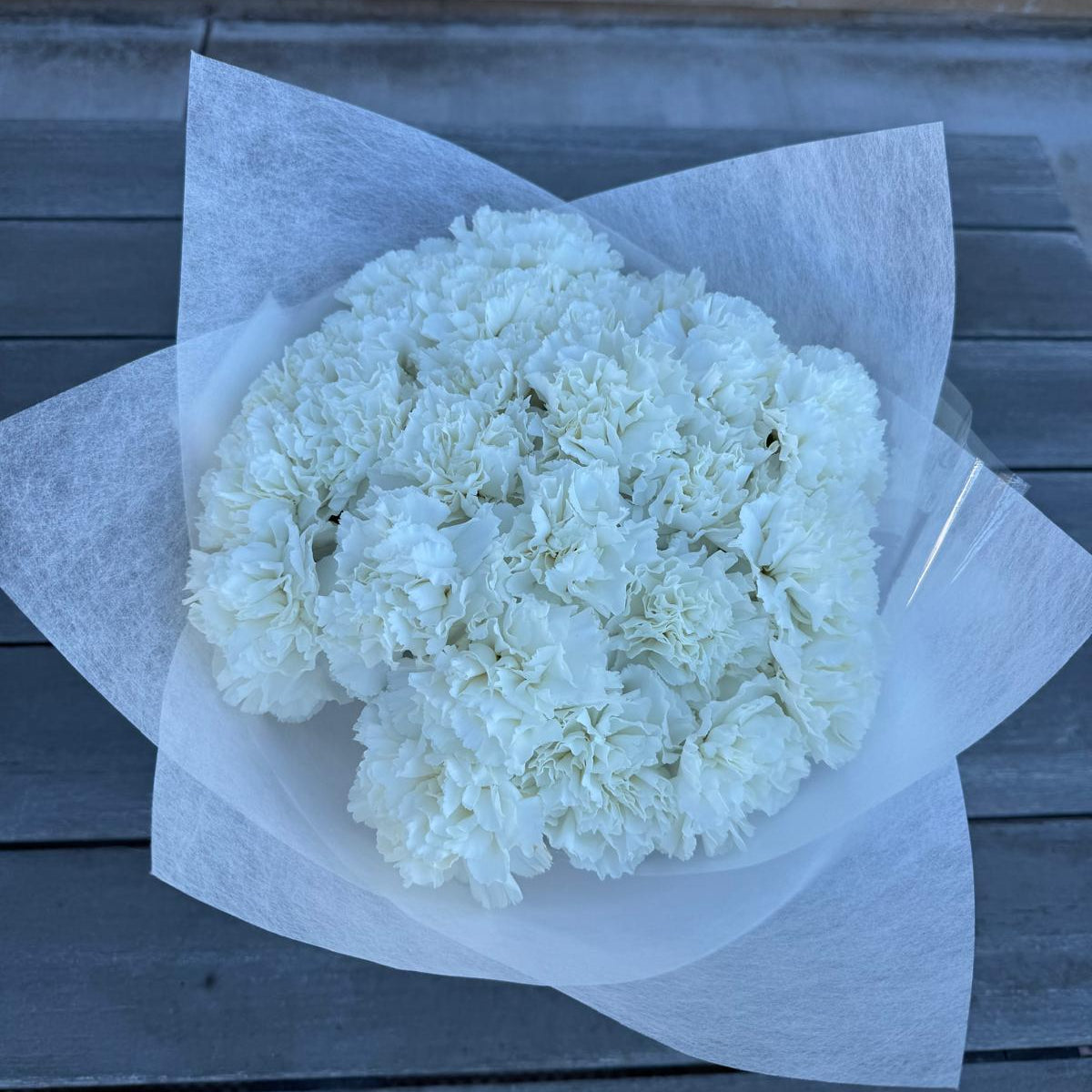 A bouquet of pure white carnations wrapped in crisp white paper, placed on a gray wooden surface. The flowers are tightly arranged, creating a soft and full appearance.