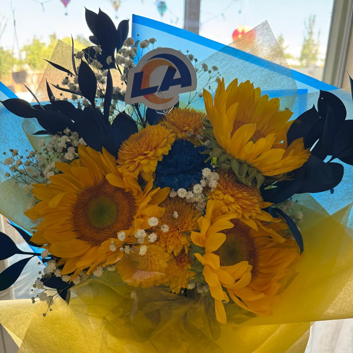 Bright bouquet of yellow sunflowers, golden chrysanthemums, blue painted leaves, and white baby’s breath. The arrangement is wrapped in yellow and blue paper with a matching yellow ribbon.