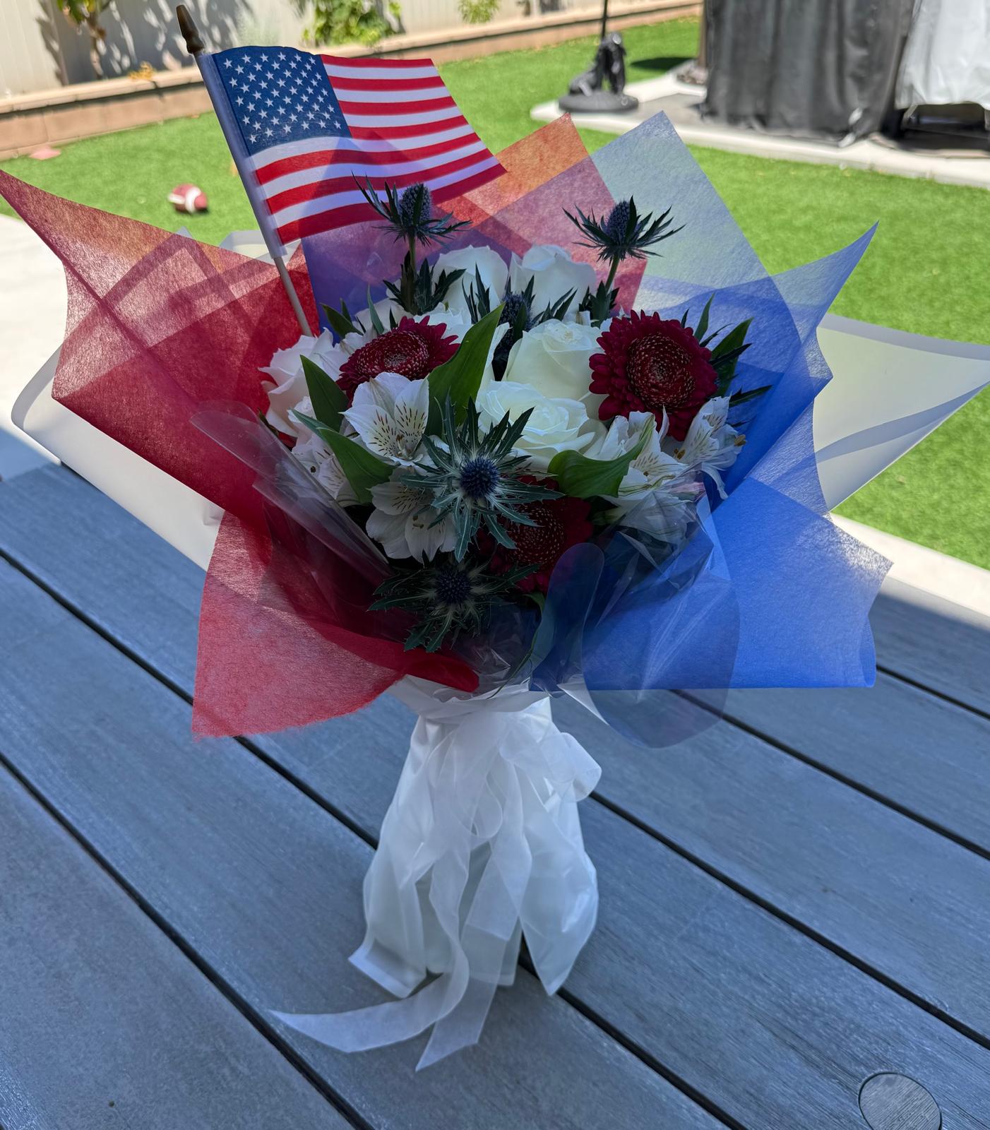 Patriotic 9/11 tribute bouquet featuring red, white, and blue flowers with thistle accents, wrapped in red and blue tissue, tied with a white ribbon, and topped with a small American flag.