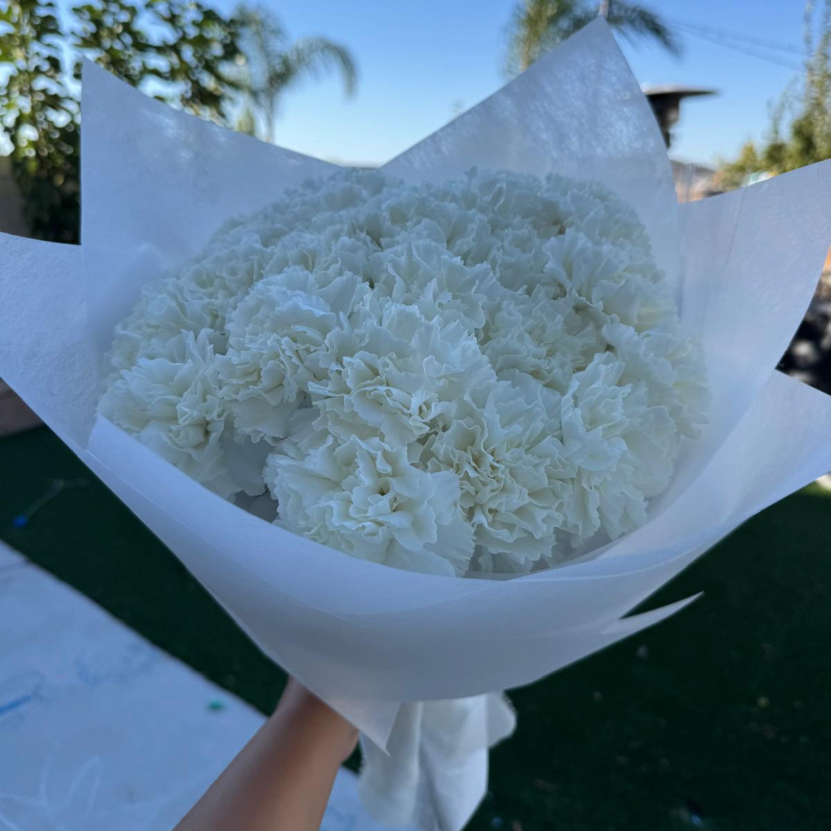 A bouquet of pure white carnations wrapped in crisp white paper, placed on a gray wooden surface. The flowers are tightly arranged, creating a soft and full appearance.