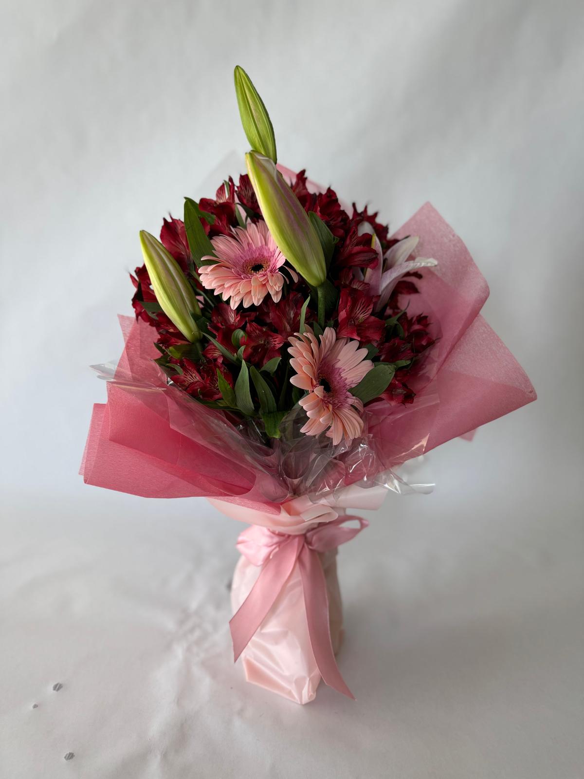 Bouquet with deep red and blush flowers including lilies, gerbera daisies and alstroemeria, wrapped in pink paper with a dramatic, romantic feel.