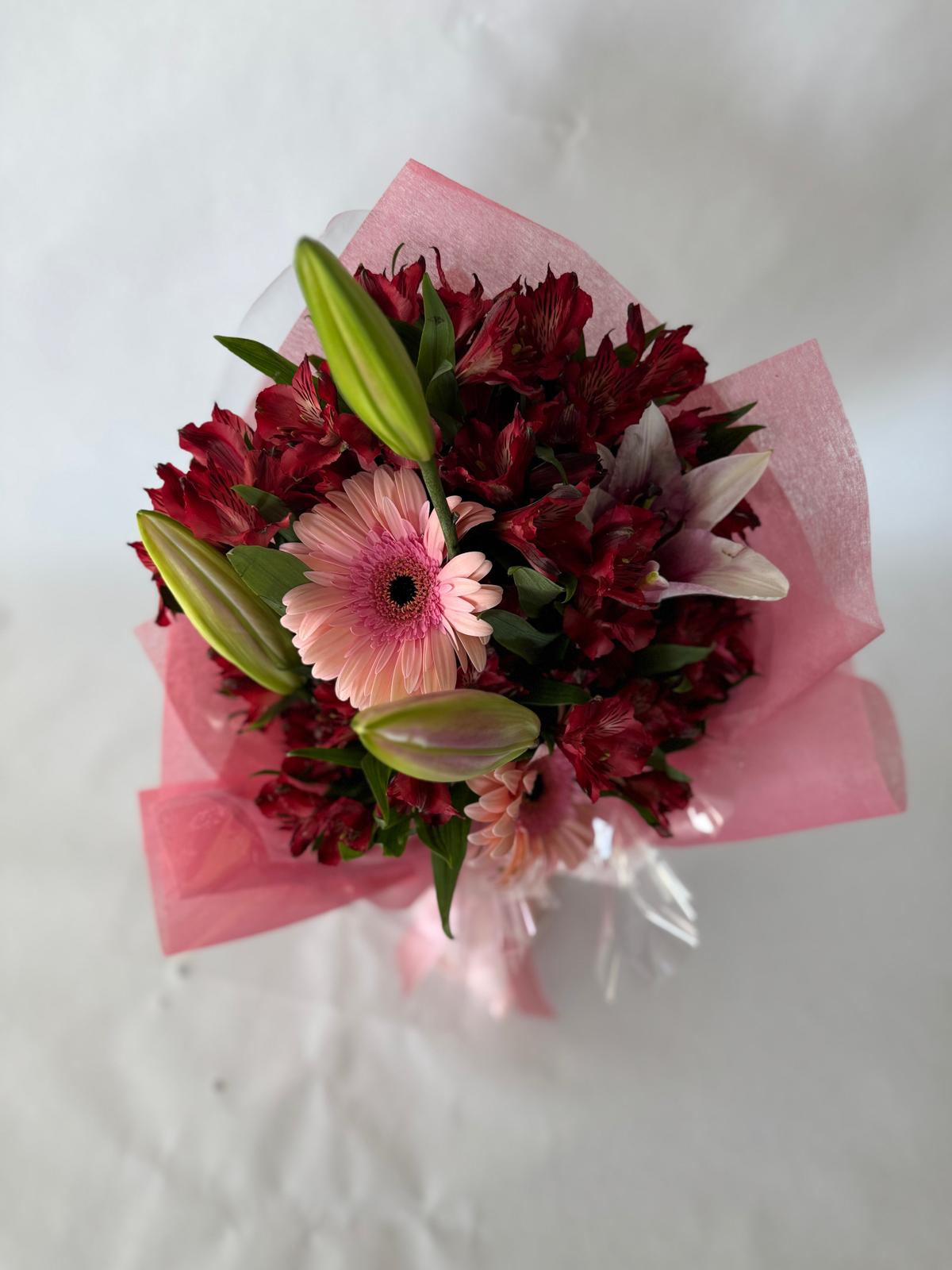 Bouquet with deep red and blush flowers including lilies, gerbera daisies and alstroemeria, wrapped in pink paper with a dramatic, romantic feel.