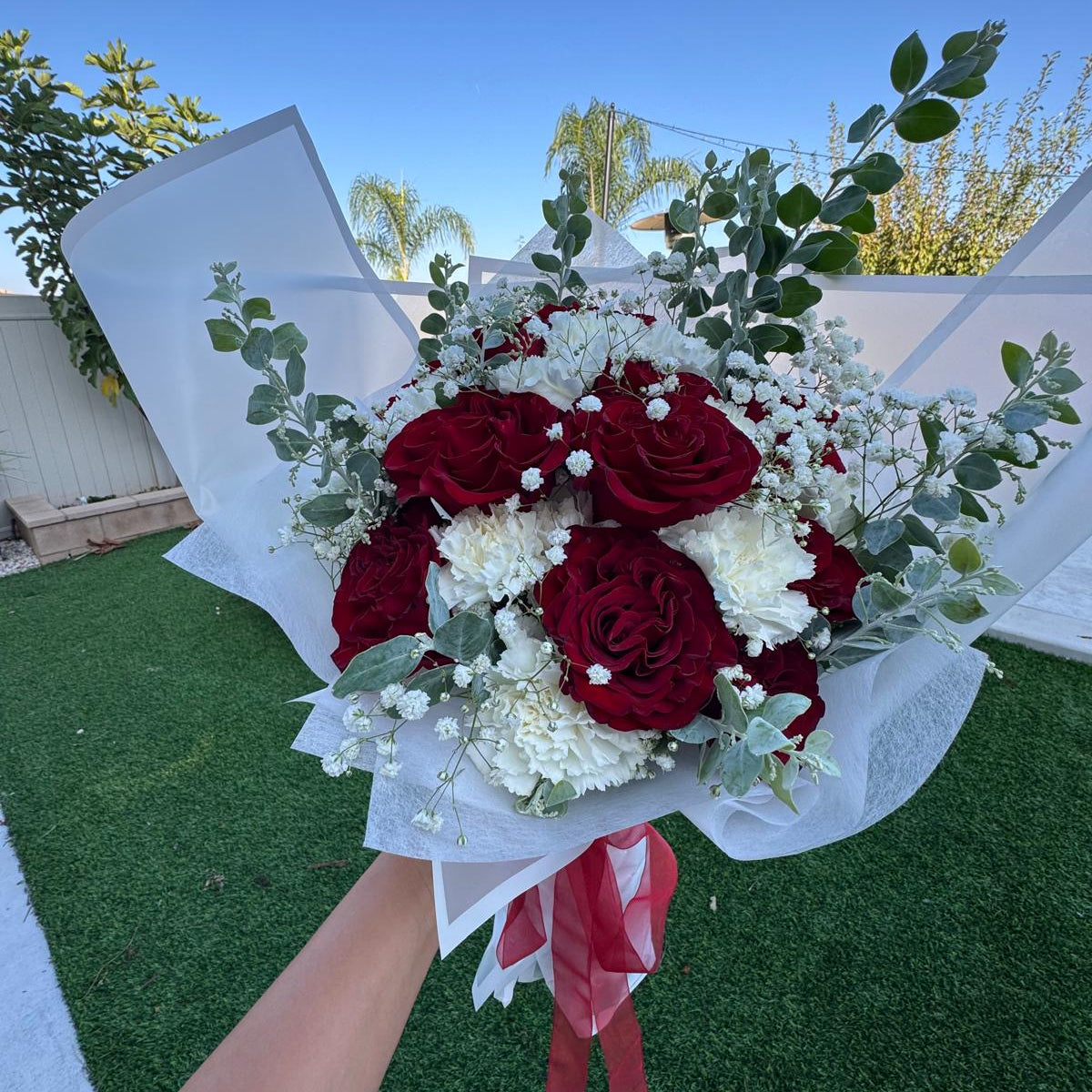 Signature Veterans Day bouquet with red roses, white carnations and baby’s breath wrapped in white paper with red ribbon by Nai Pascoal Bouquets.