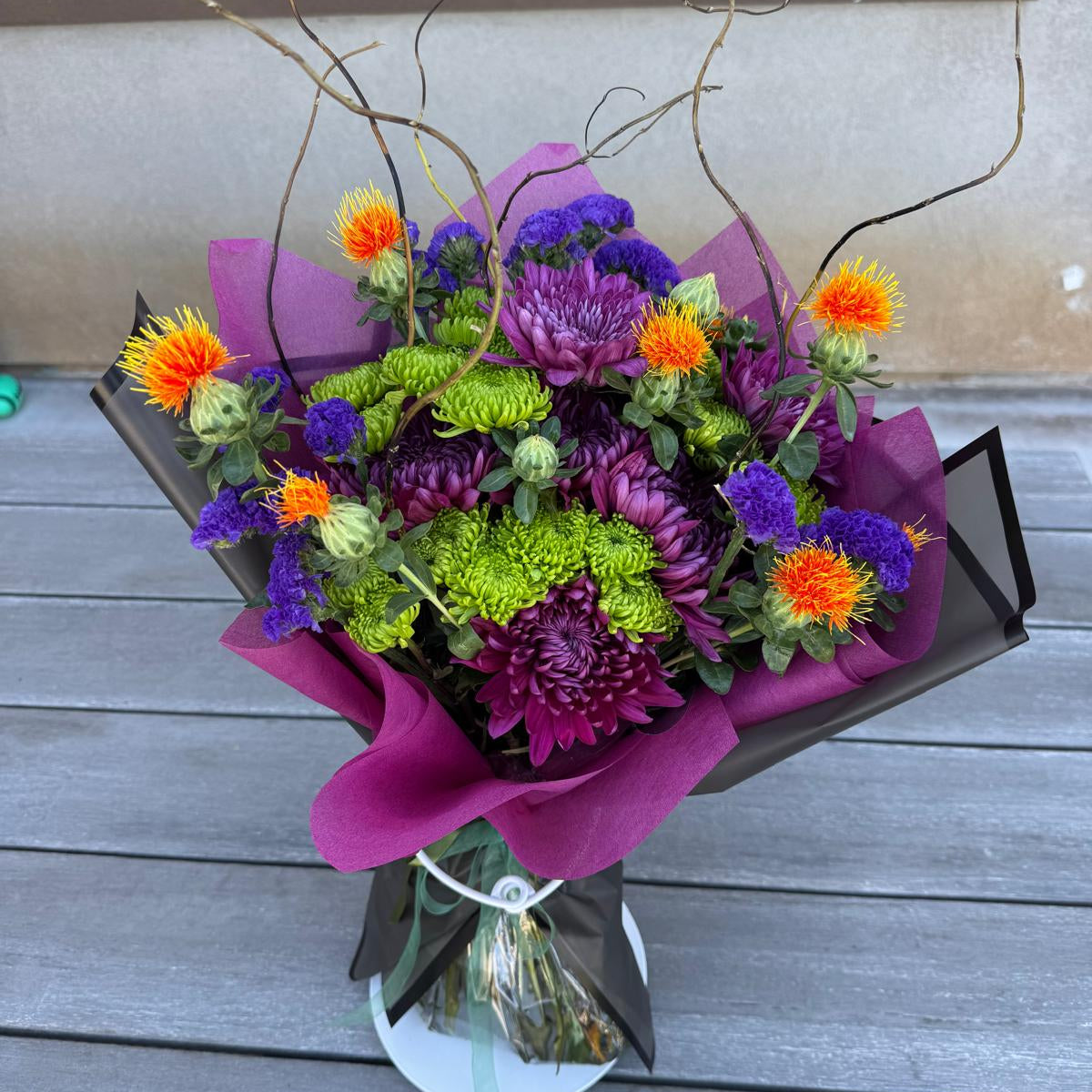 Witch’s Brew Bouquet featuring purple, orange, and green flowers with curly willow branches, wrapped in black.