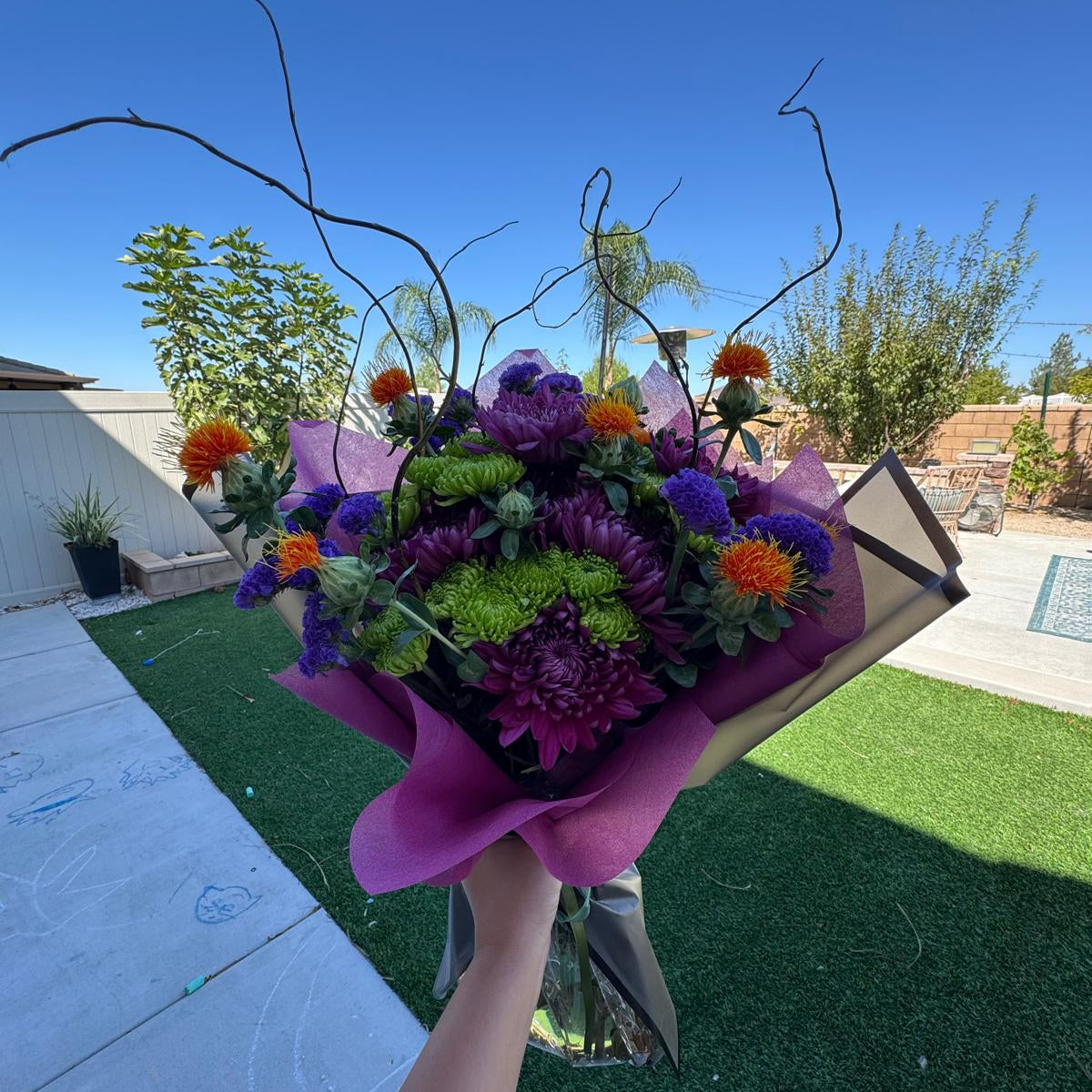 Witch’s Brew Bouquet featuring purple, orange, and green flowers with curly willow branches, wrapped in black.