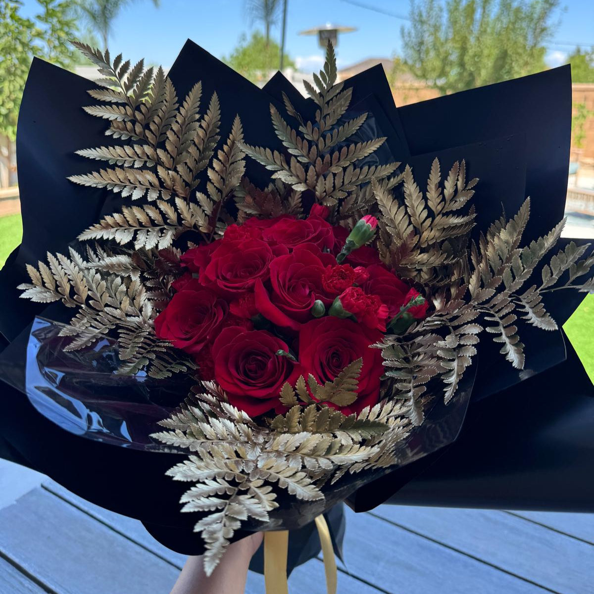 Full view of the 49ers-inspired bouquet, with rich red flowers framed by golden ferns, wrapped in glossy black paper and tied with a metallic gold ribbon.