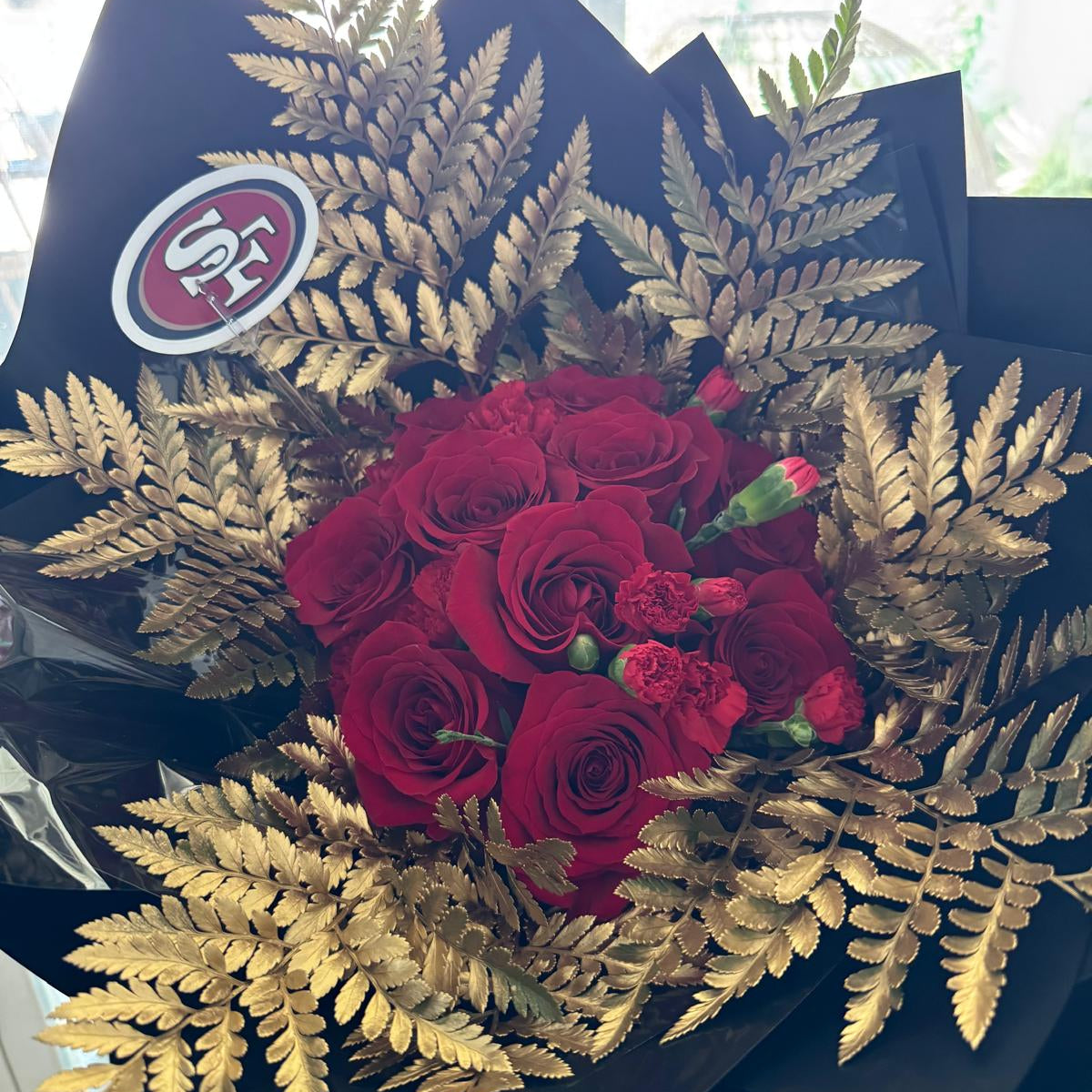 Full view of the 49ers-inspired bouquet, with rich red flowers framed by golden ferns, wrapped in glossy black paper and tied with a metallic gold ribbon.