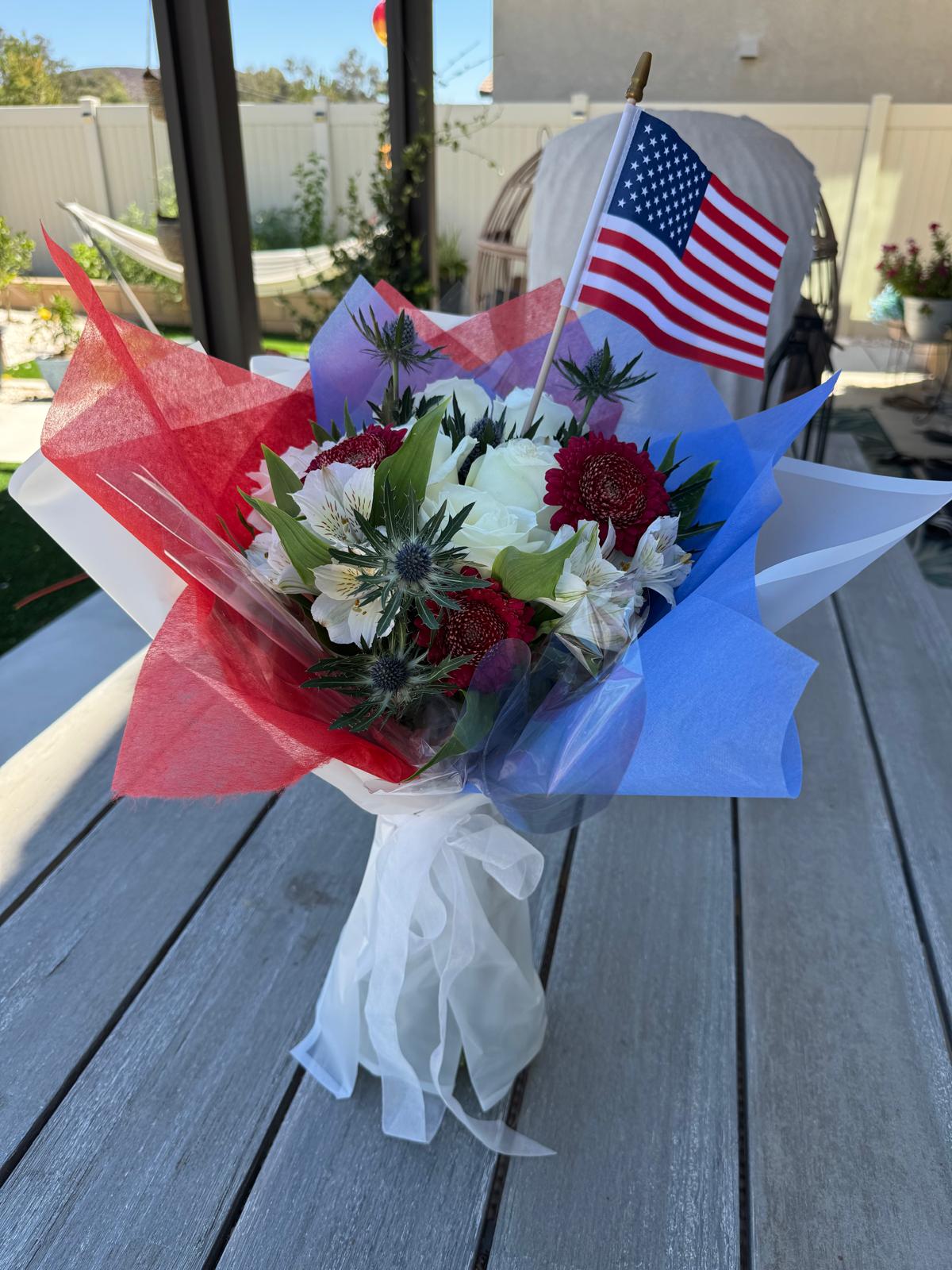Patriotic 9/11 tribute bouquet featuring red, white, and blue flowers with thistle accents, wrapped in red and blue tissue, tied with a white ribbon, and topped with a small American flag.