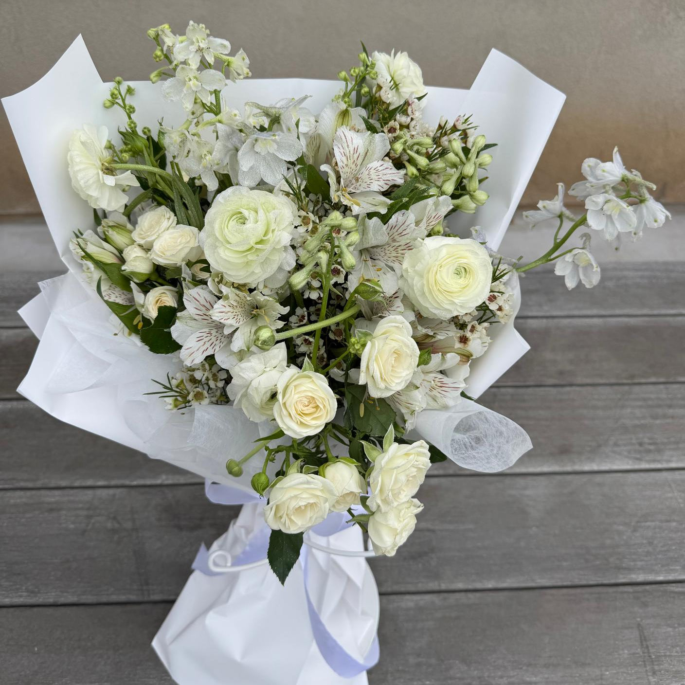 April Diamond bouquet featuring soft white roses, ranunculus, and delicate blooms, elegantly wrapped in white paper with a ribbon, styled on a wooden backdrop.