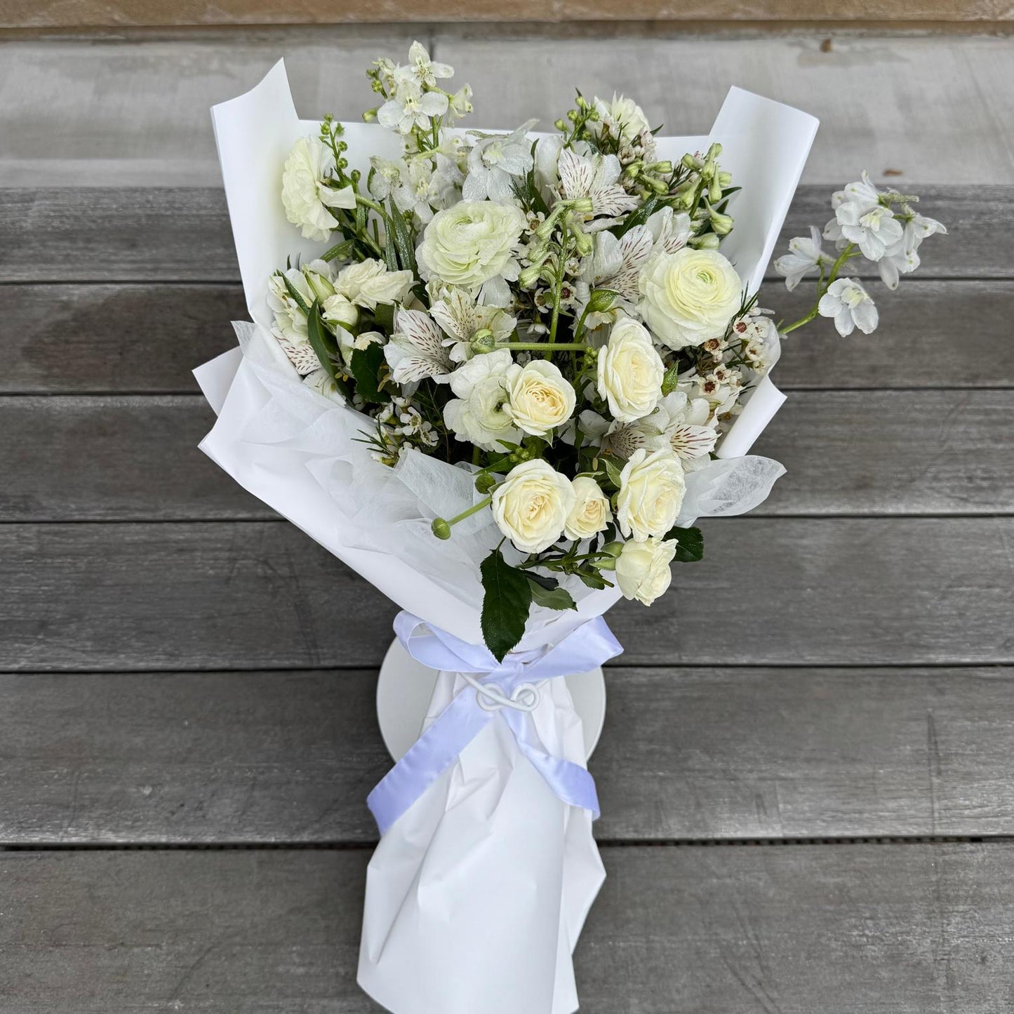 April Diamond bouquet featuring soft white roses, ranunculus, and delicate blooms, elegantly wrapped in white paper with a ribbon, styled on a wooden backdrop.