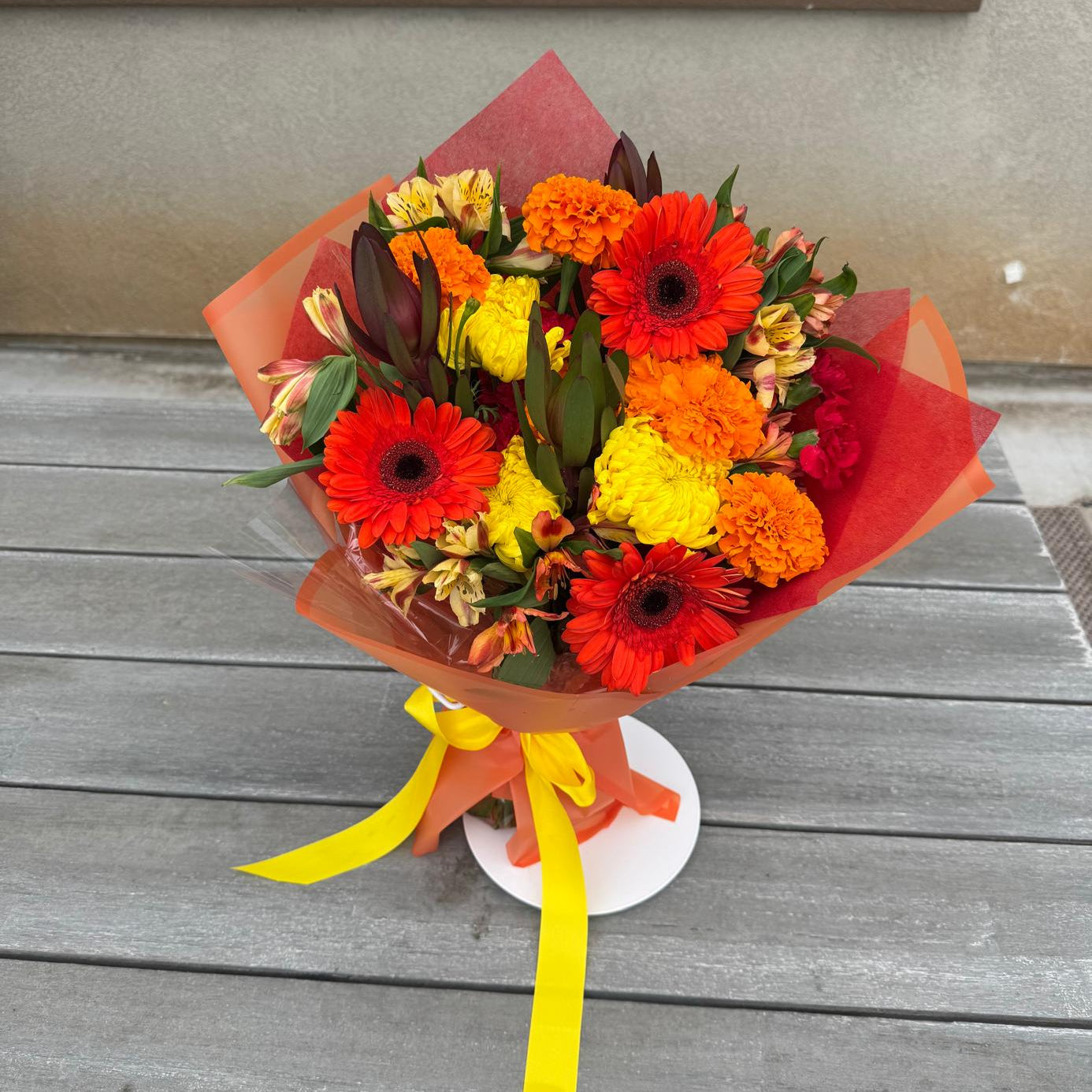 Bright fall bouquet with orange gerbera daisies, yellow mums, marigolds, and alstroemeria wrapped in autumn-colored paper with a yellow ribbon.