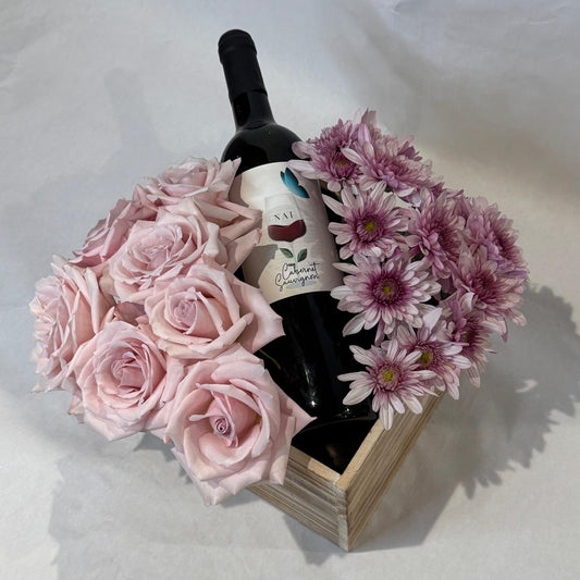 A floral gift box featuring blush roses and lavender mums arranged around a wine bottle in a wooden crate. Wine not included.