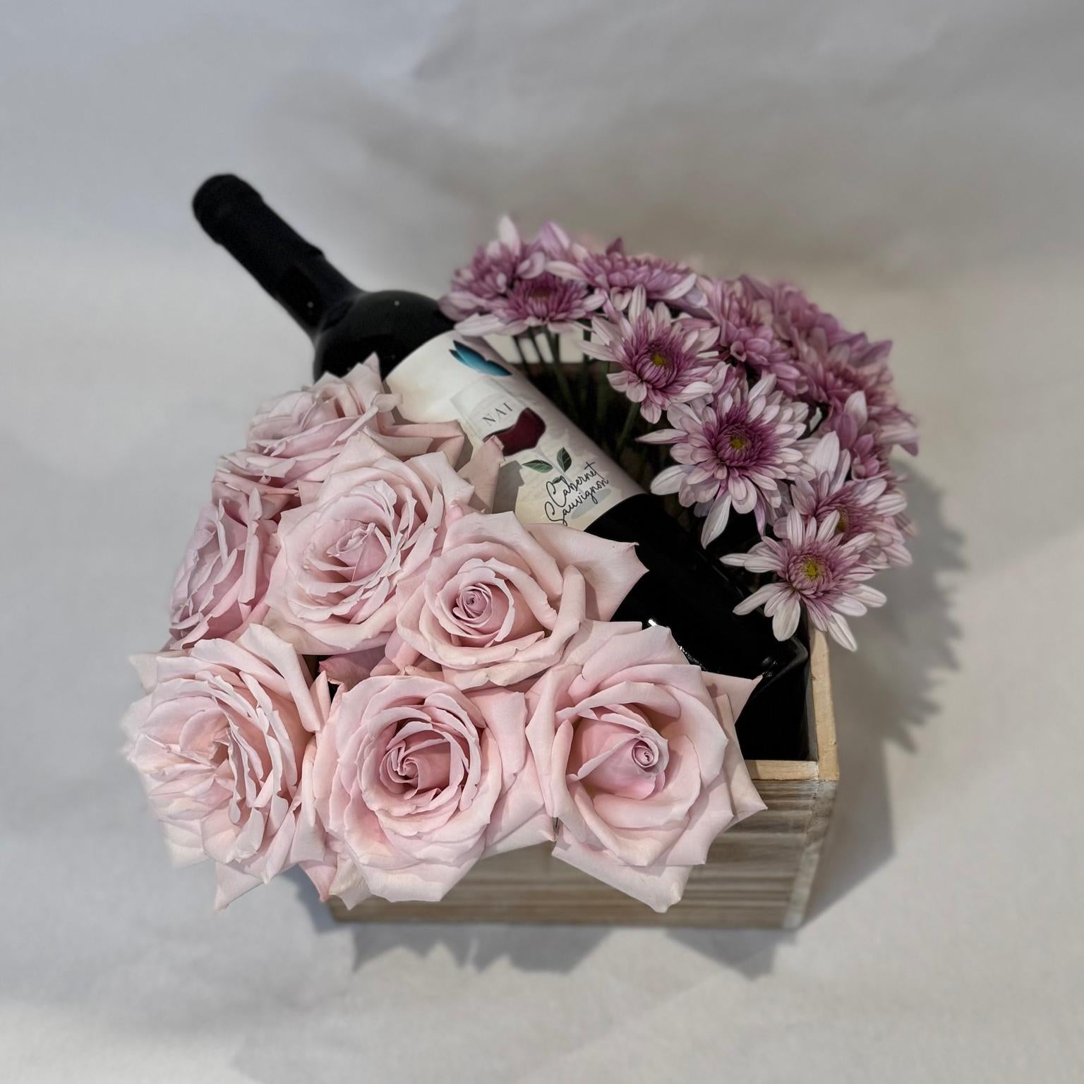 A floral gift box featuring blush roses and lavender mums arranged around a wine bottle in a wooden crate. Wine not included.
