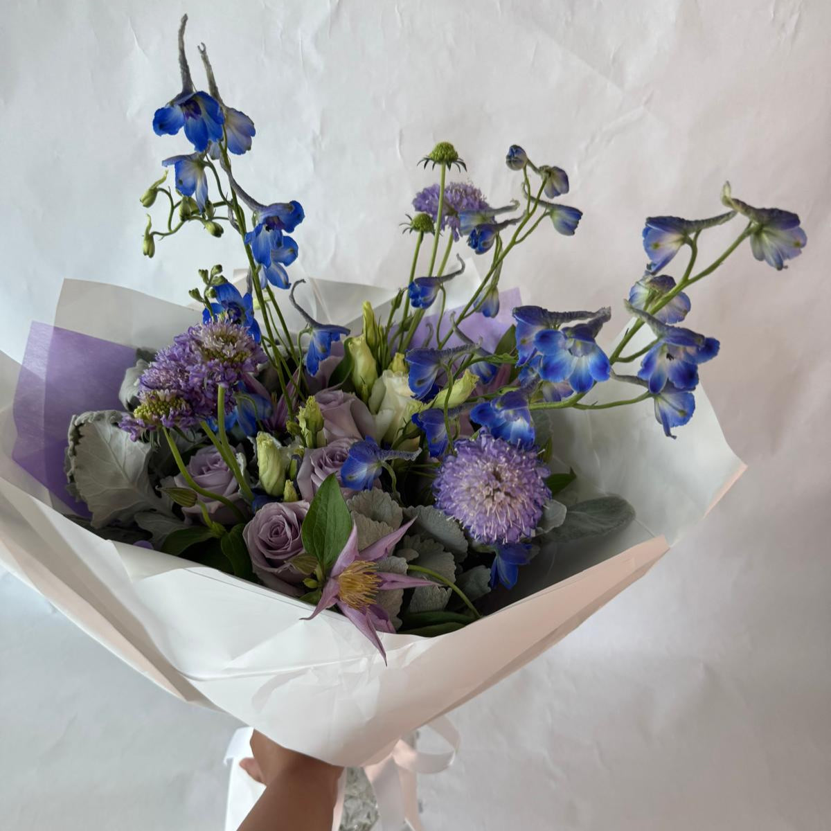 A soothing bouquet in calming shades of lavender, blue and white, beautifully wrapped in lilac and white paper with a white ribbon, designed to evoke peace, healing and quiet strength.