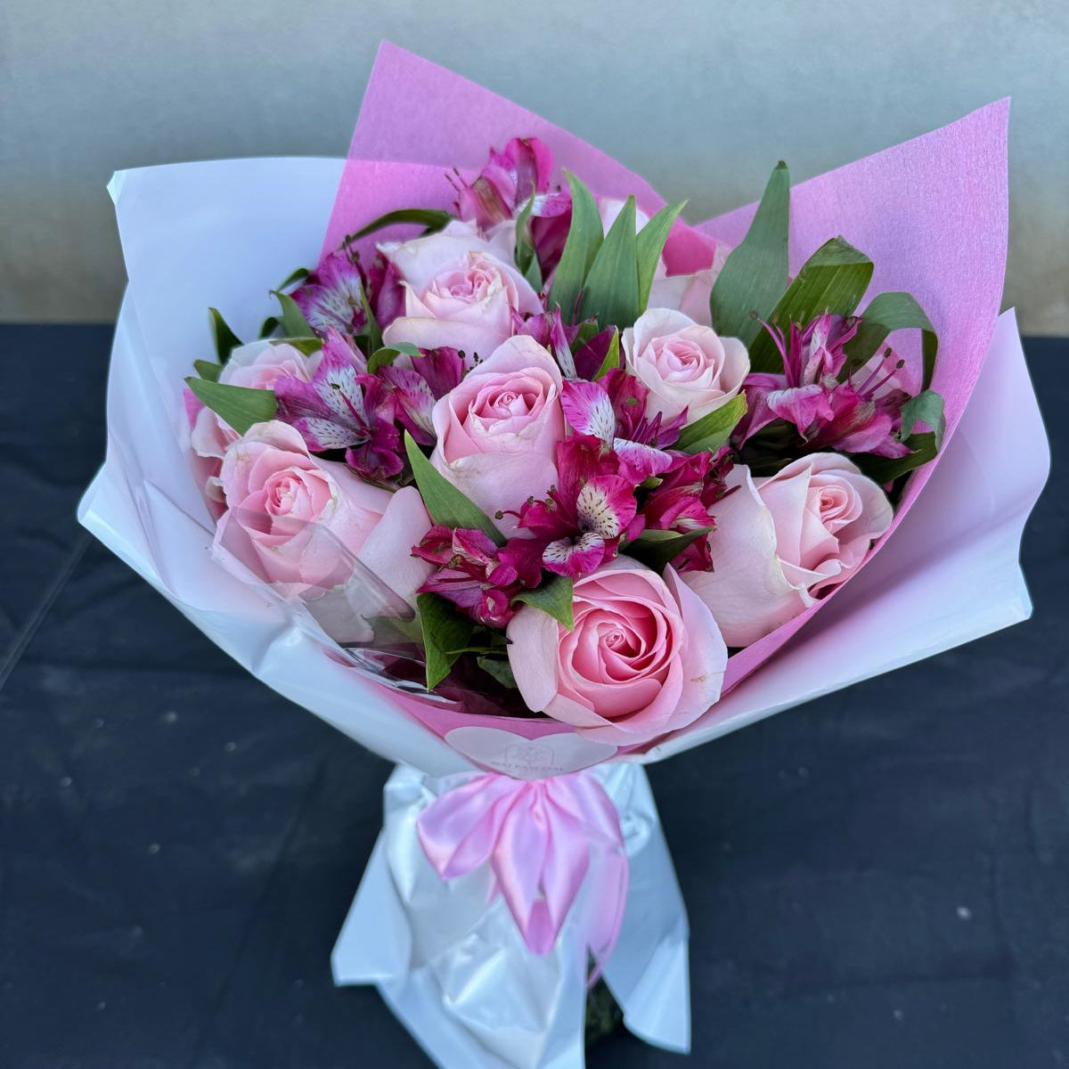 Blushing Grace Bouquet in one size only featuring blush pink roses and magenta alstroemeria wrapped in pink and white paper with satin ribbon by Nai Pascoal Bouquets
