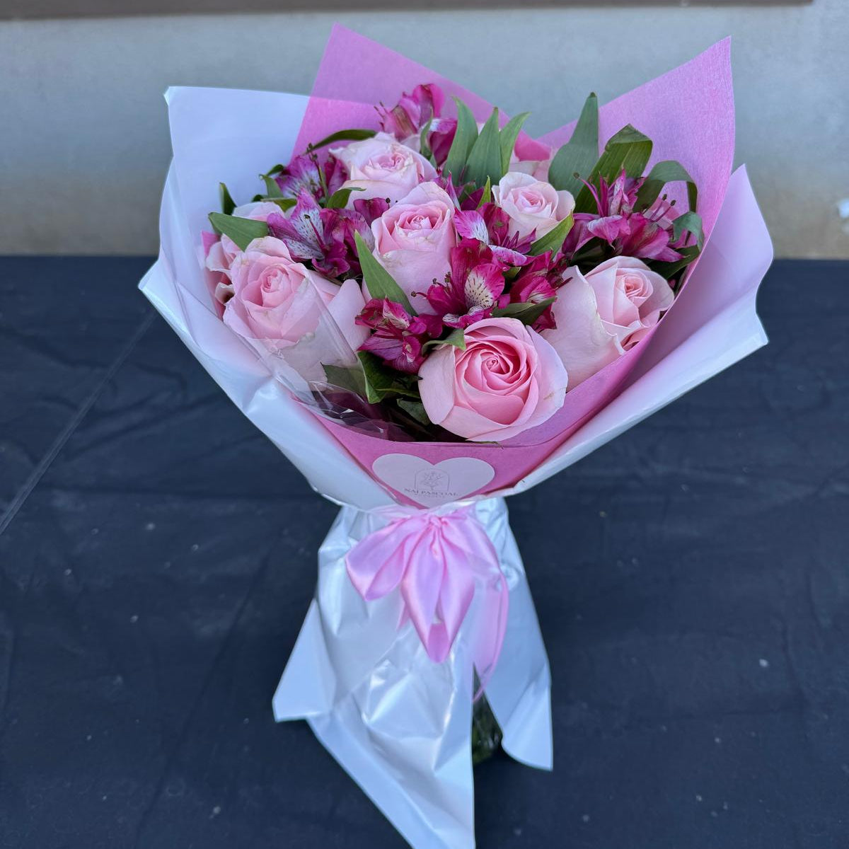 Blushing Grace Bouquet in one size only featuring blush pink roses and magenta alstroemeria wrapped in pink and white paper with satin ribbon by Nai Pascoal Bouquets
