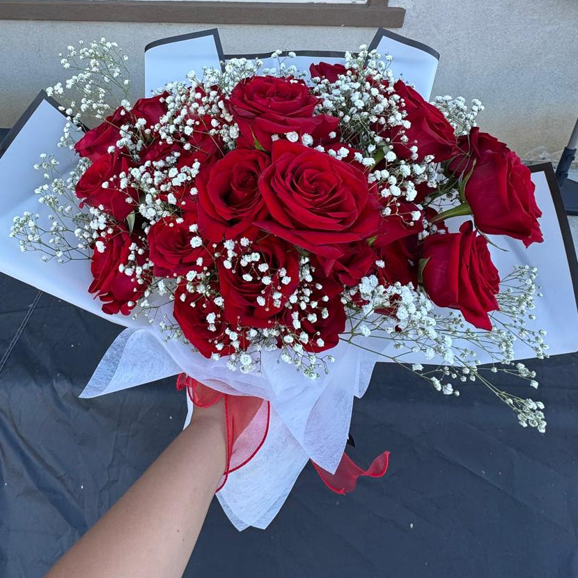 Hand-tied bouquet of red roses with white baby’s breath, wrapped in white paper with a red ribbon.