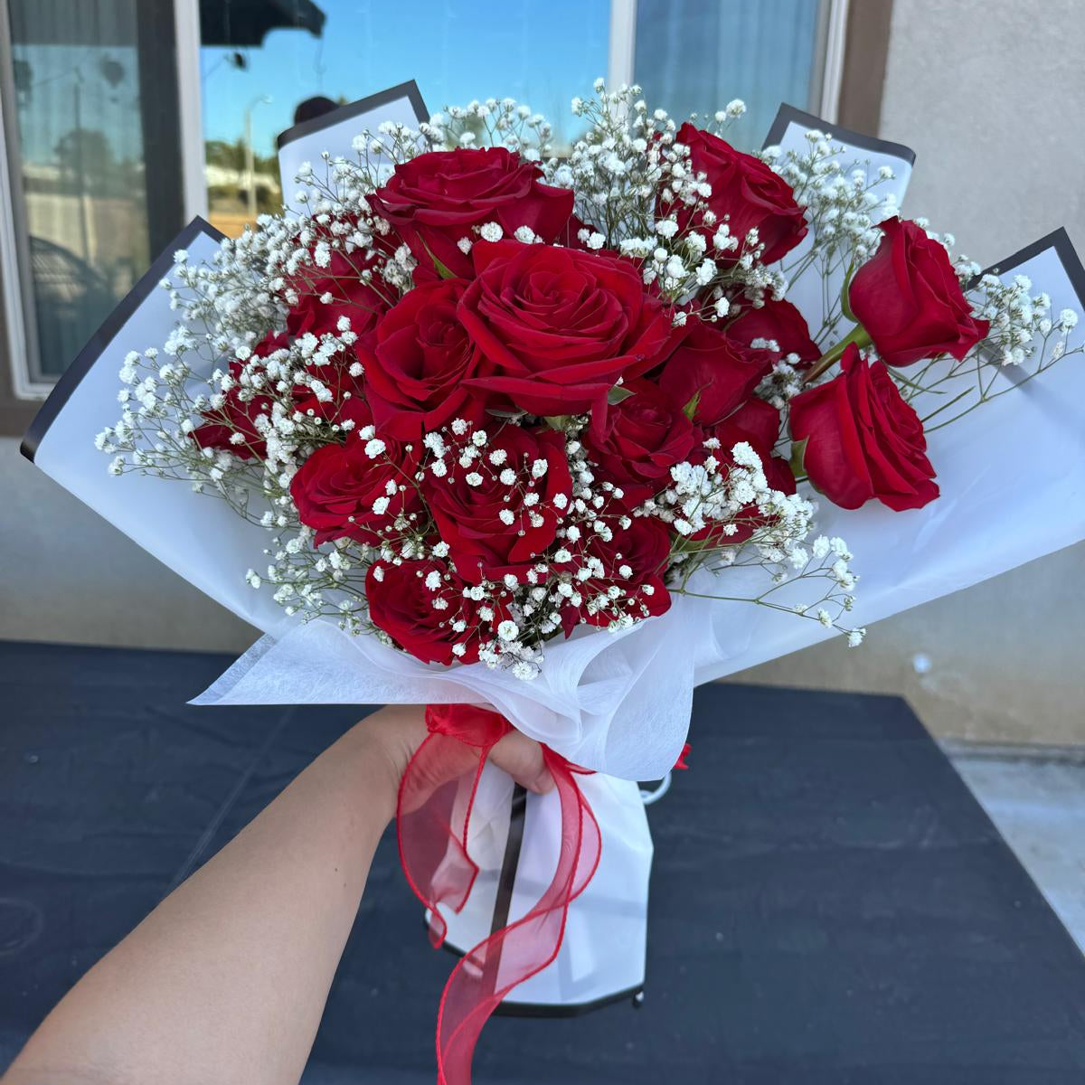 Hand-tied bouquet of red roses with white baby’s breath, wrapped in white paper with a red ribbon.