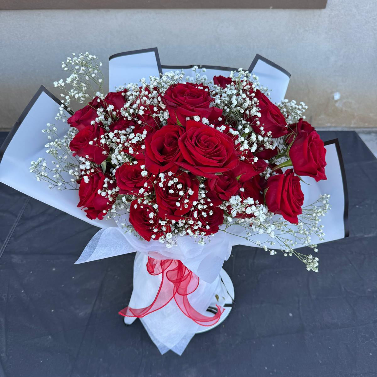 Hand-tied bouquet of red roses with white baby’s breath, wrapped in white paper with a red ribbon.