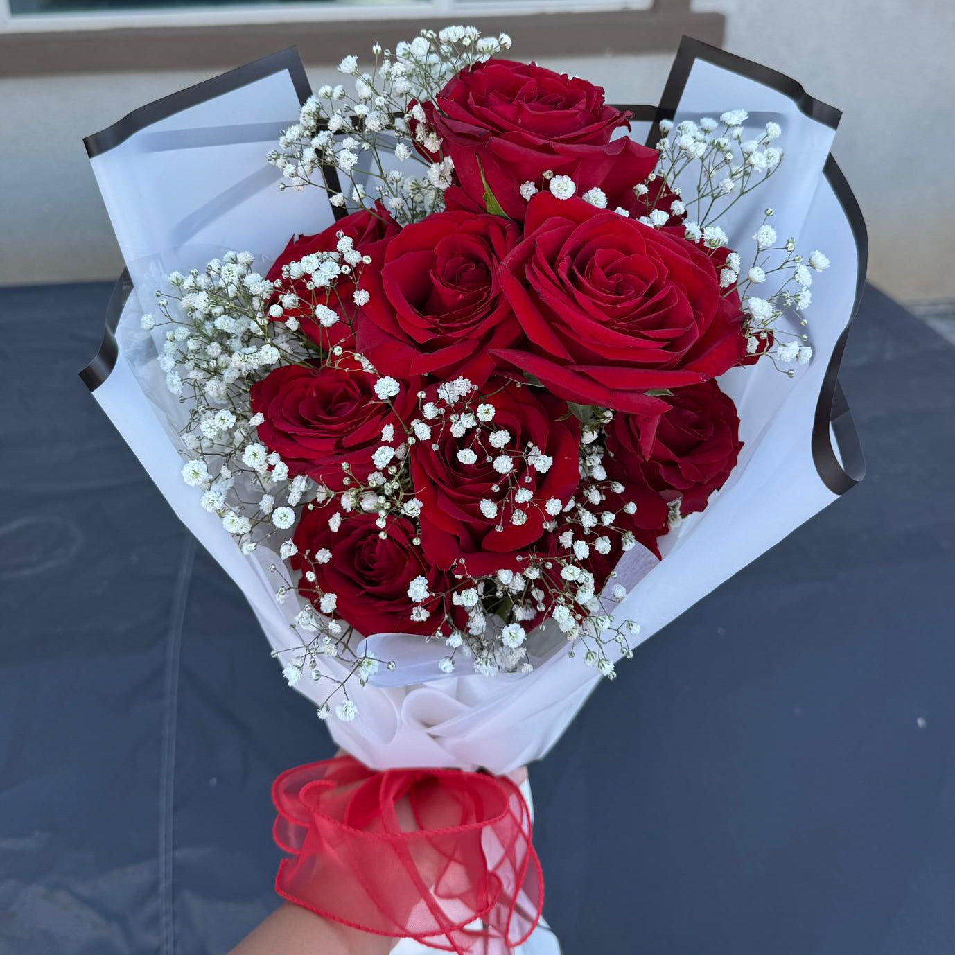 Hand-tied bouquet of red roses with white baby’s breath, wrapped in white paper with a red ribbon.