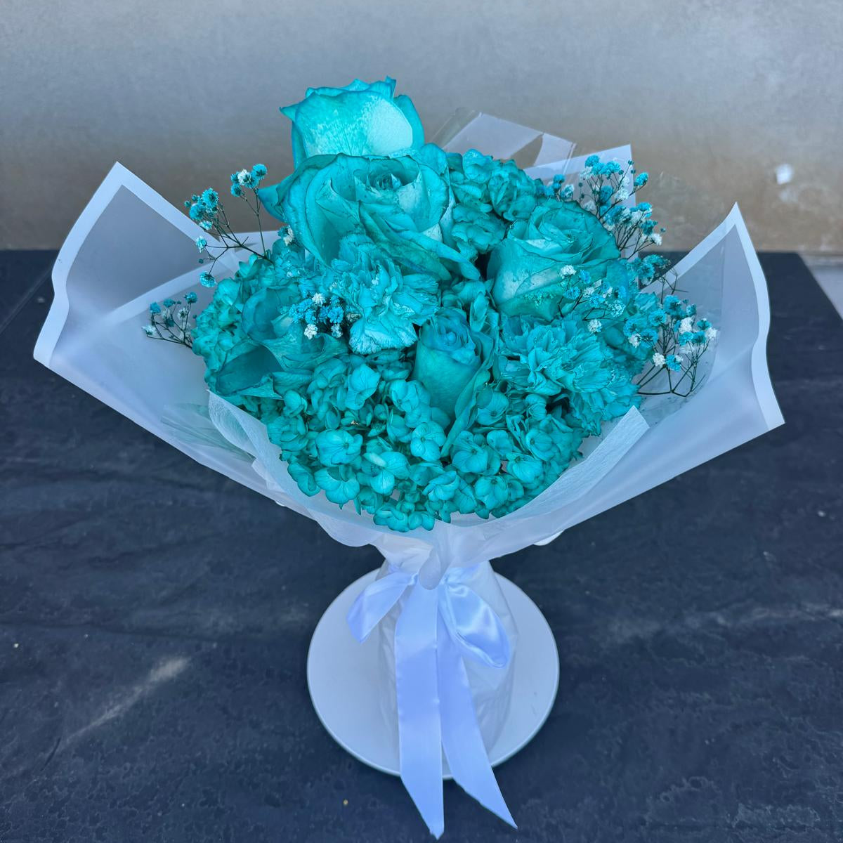 A monochromatic turquoise bouquet featuring fully turquoise-dyed blooms arranged tightly in a round, dome-style shape. The bouquet is wrapped in crisp white translucent paper and tied with a white satin ribbon, displayed on a white stand against a dark surface.
