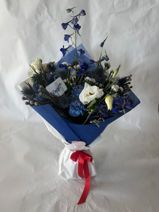Signature floral bouquet in Dodgers team colors, wrapped in Korean paper and tied with a silk ribbon. Features blue, white, and soft neutral blooms with matching team sticker