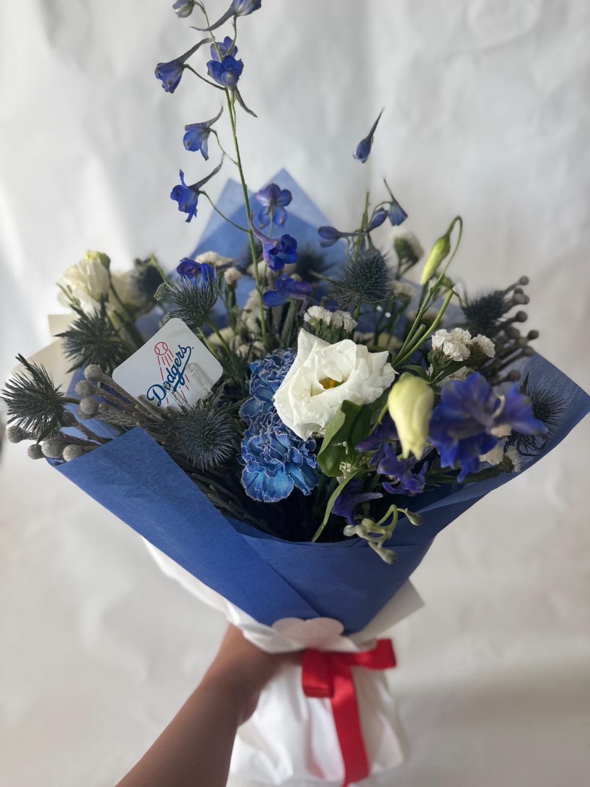 Signature floral bouquet in Dodgers team colors, wrapped in Korean paper and tied with a silk ribbon. Features blue, white, and soft neutral blooms with matching team sticker
