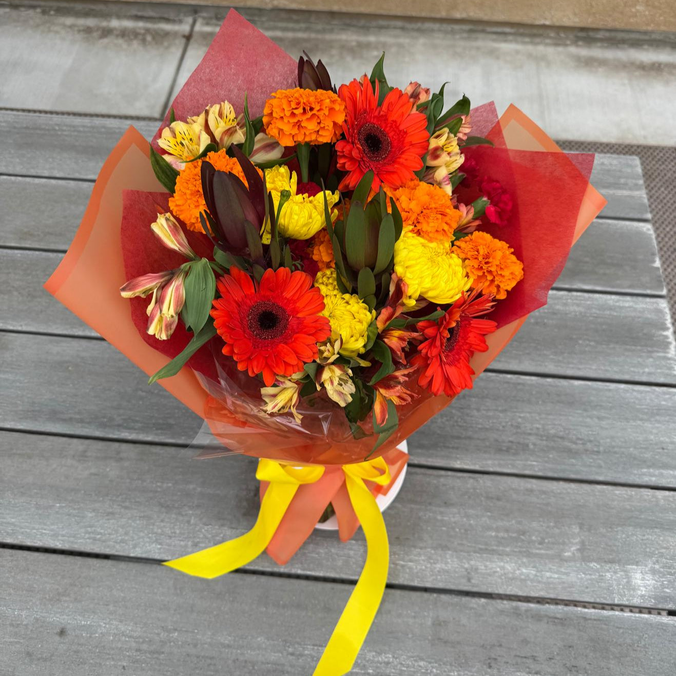 Bright fall bouquet with orange gerbera daisies, yellow mums, marigolds, and alstroemeria wrapped in autumn-colored paper with a yellow ribbon.