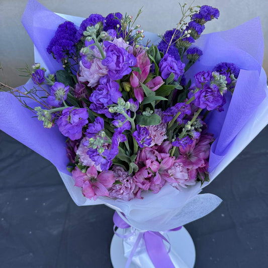 Purple and lavender February bouquet featuring stock, carnations, alstroemeria, and statice wrapped in soft lavender paper.