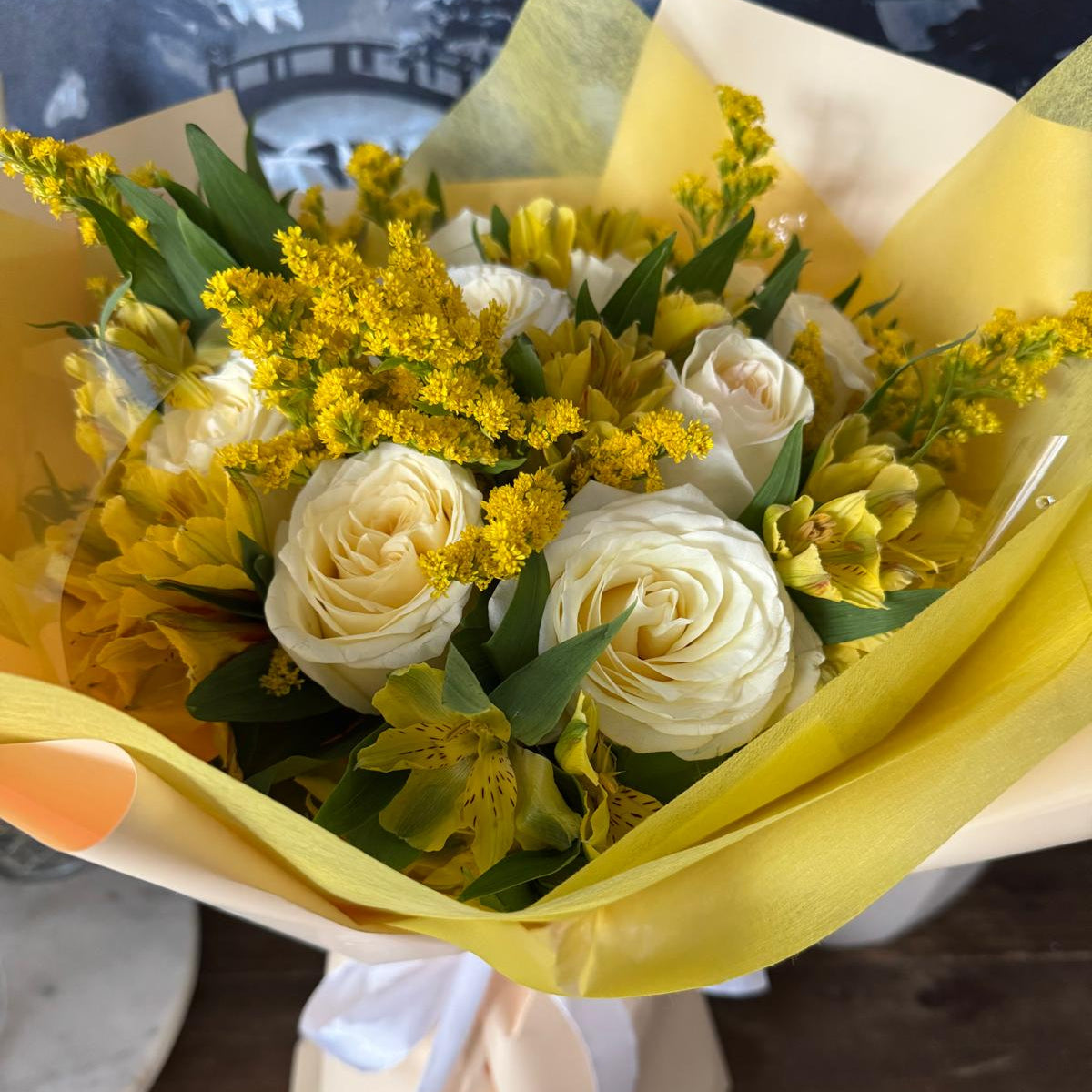 Golden Daylight Bouquet in one size only featuring creamy white roses, yellow alstroemeria, and golden accents wrapped in yellow paper by Nai Pascoal Bouquets