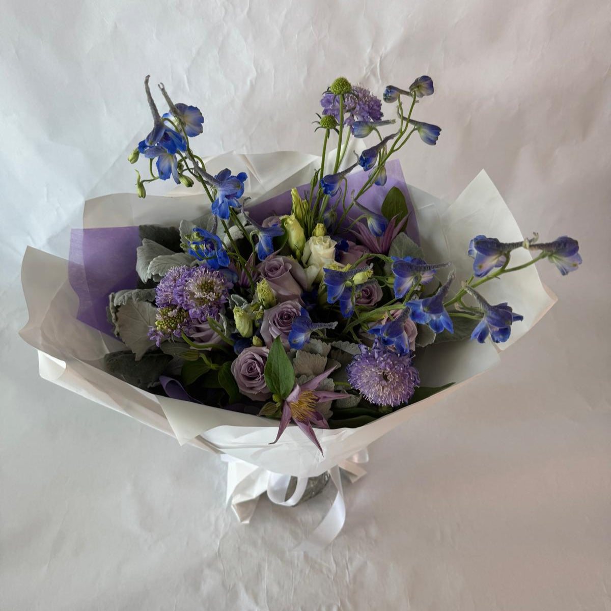 A soothing bouquet in calming shades of lavender, blue and white, beautifully wrapped in lilac and white paper with a white ribbon, designed to evoke peace, healing and quiet strength.