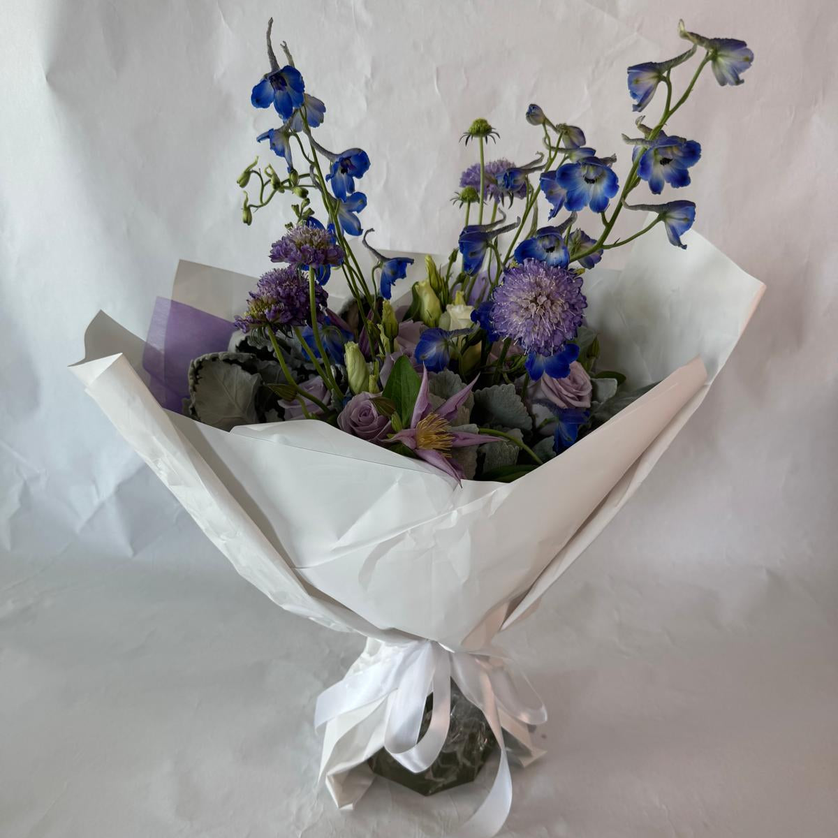A soothing bouquet in calming shades of lavender, blue and white, beautifully wrapped in lilac and white paper with a white ribbon, designed to evoke peace, healing and quiet strength.