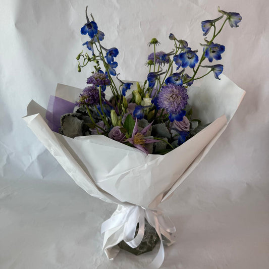 A soothing bouquet in calming shades of lavender, blue and white, beautifully wrapped in lilac and white paper with a white ribbon, designed to evoke peace, healing and quiet strength.