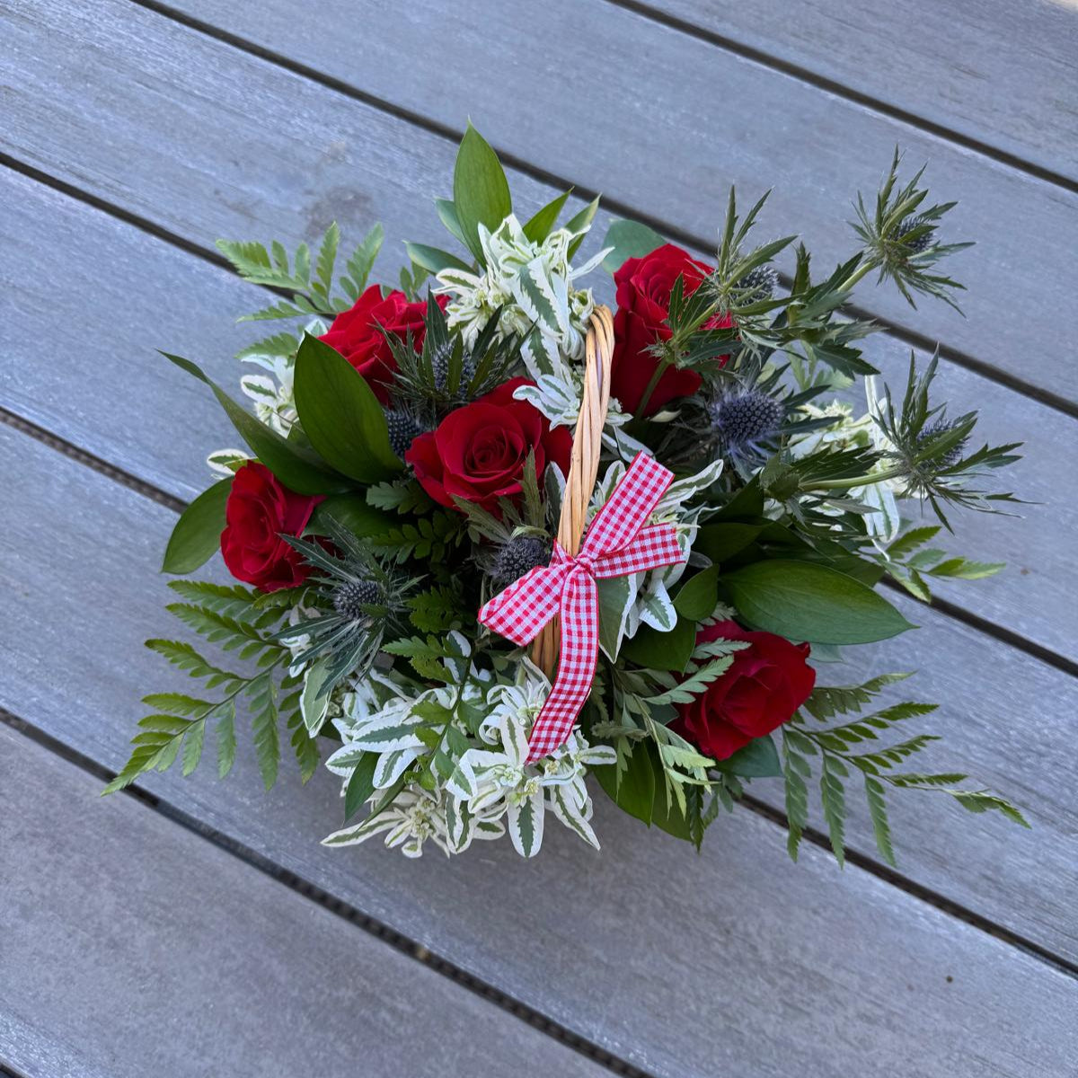 Festive Labor Day flower basket with vibrant red roses, white variegated foliage, green ferns, and thistle accents, finished with a red-and-white gingham bow.