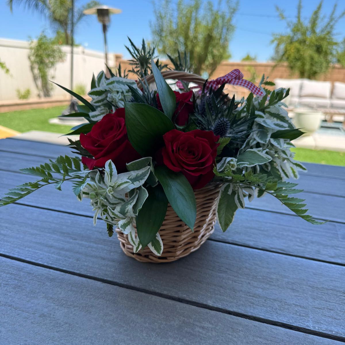 Festive Labor Day flower basket with vibrant red roses, white variegated foliage, green ferns, and thistle accents, finished with a red-and-white gingham bow.