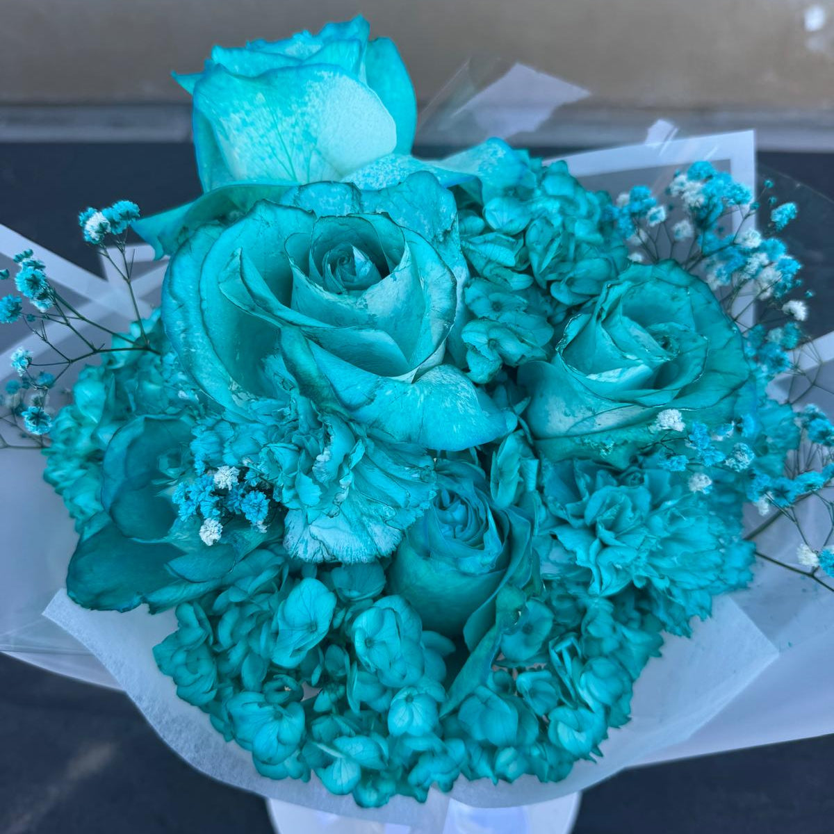 A monochromatic turquoise bouquet featuring fully turquoise-dyed blooms arranged tightly in a round, dome-style shape. The bouquet is wrapped in crisp white translucent paper and tied with a white satin ribbon, displayed on a white stand against a dark surface.