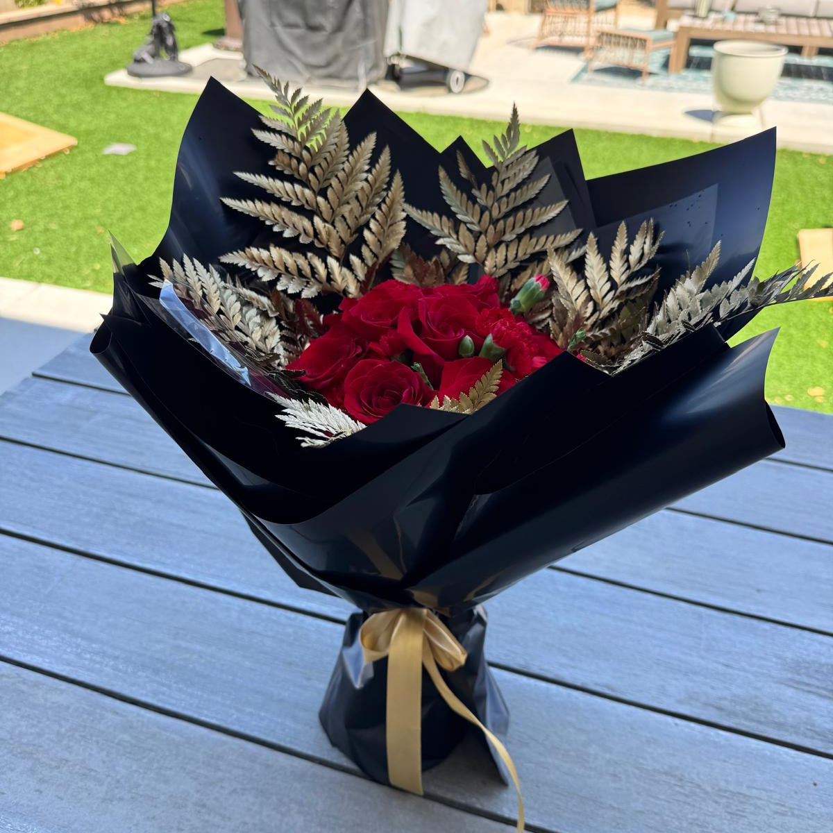 Full view of the 49ers-inspired bouquet, with rich red flowers framed by golden ferns, wrapped in glossy black paper and tied with a metallic gold ribbon.