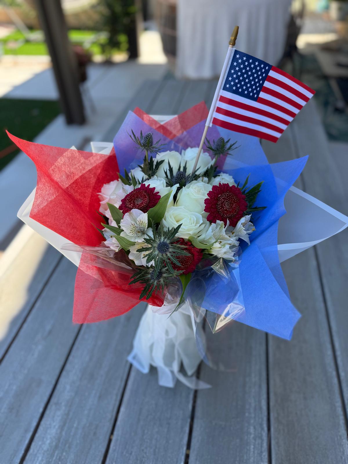 Patriotic 9/11 tribute bouquet featuring red, white, and blue flowers with thistle accents, wrapped in red and blue tissue, tied with a white ribbon, and topped with a small American flag.