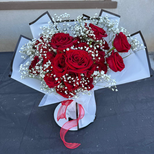 Hand-tied bouquet of red roses with white baby’s breath, wrapped in white paper with a red ribbon.