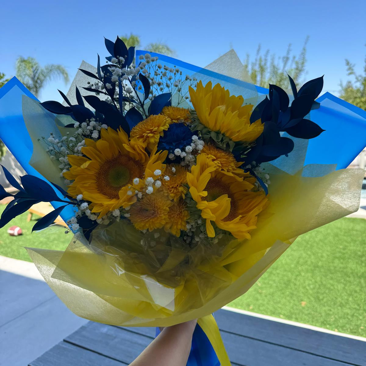 Bright bouquet of yellow sunflowers, golden chrysanthemums, blue painted leaves, and white baby’s breath. The arrangement is wrapped in yellow and blue paper with a matching yellow ribbon.
