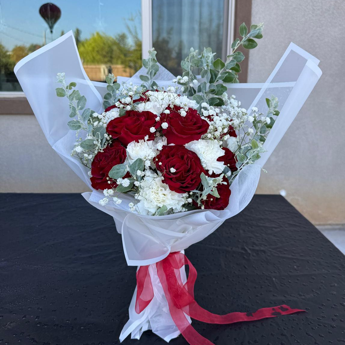 Signature Veterans Day bouquet with red roses, white carnations and baby’s breath wrapped in white paper with red ribbon by Nai Pascoal Bouquets.