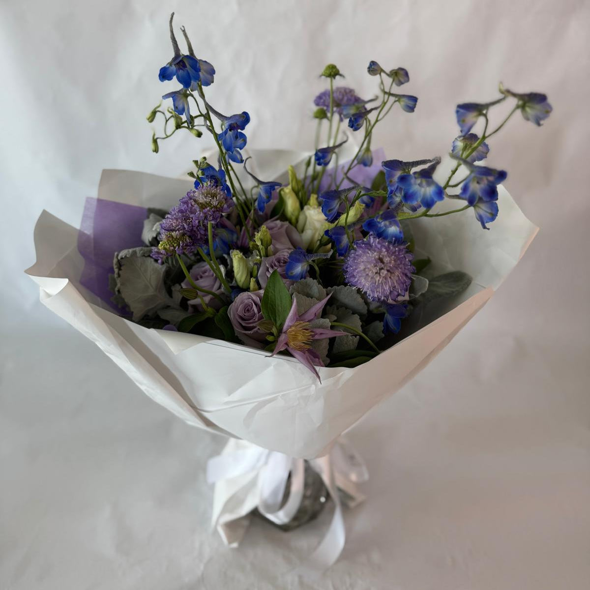 A soothing bouquet in calming shades of lavender, blue and white, beautifully wrapped in lilac and white paper with a white ribbon, designed to evoke peace, healing and quiet strength.