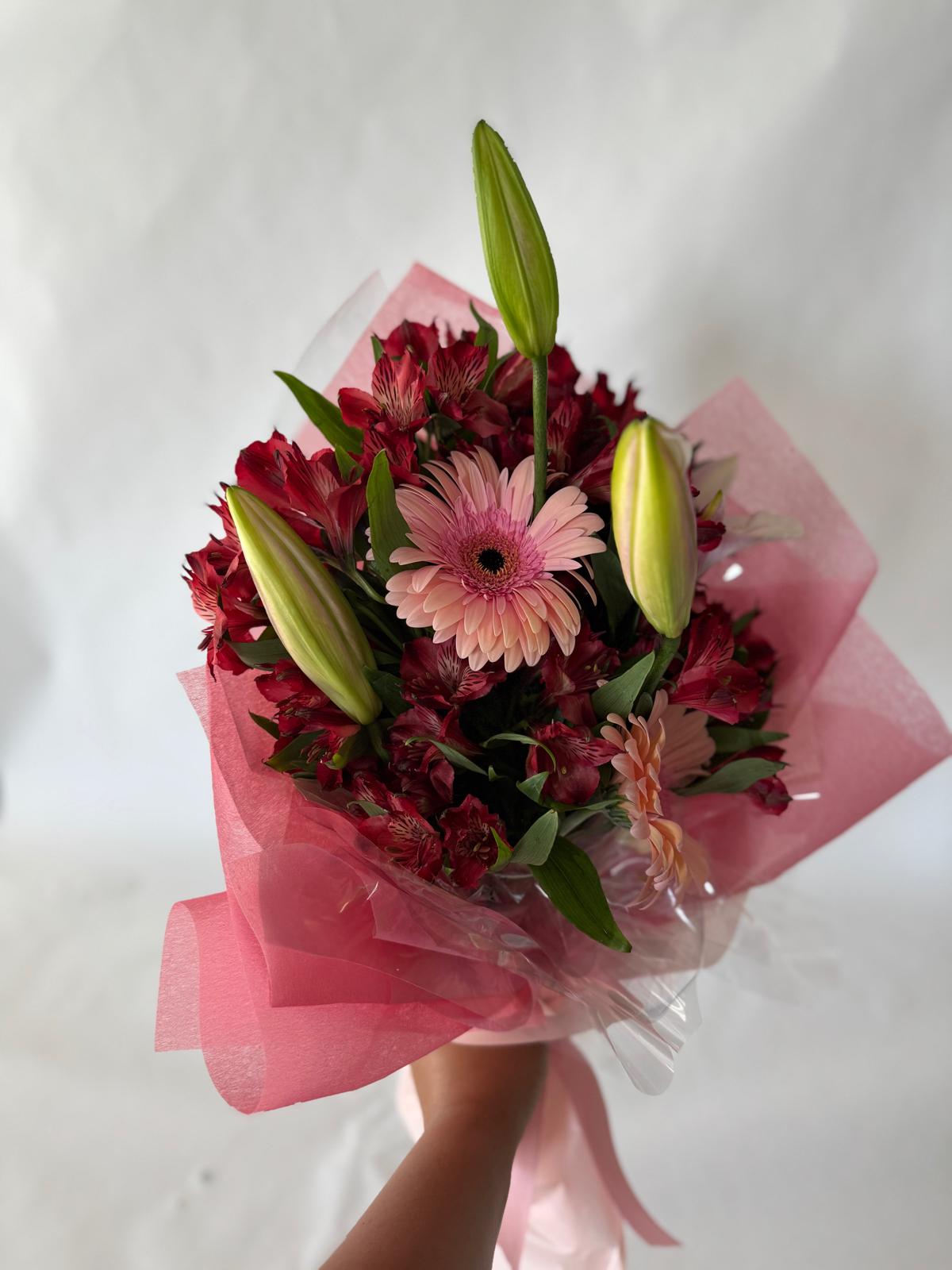 Bouquet with deep red and blush flowers including lilies, gerbera daisies and alstroemeria, wrapped in pink paper with a dramatic, romantic feel.