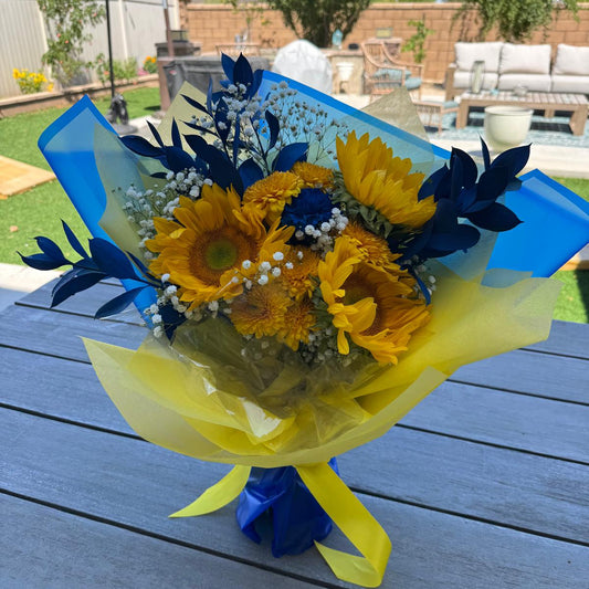 Bright bouquet of yellow sunflowers, golden chrysanthemums, blue painted leaves, and white baby’s breath. The arrangement is wrapped in yellow and blue paper with a matching yellow ribbon.