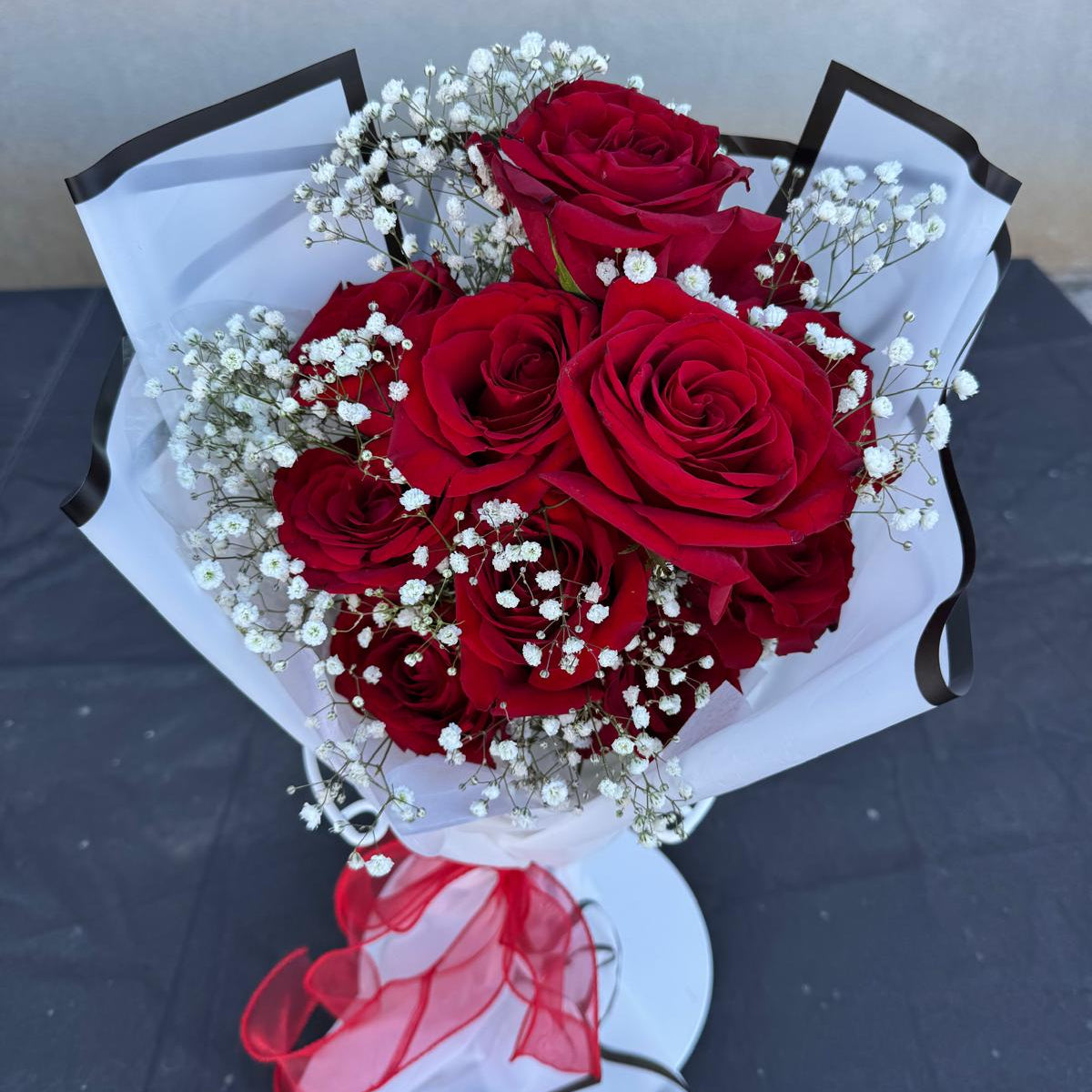 Hand-tied bouquet of red roses with white baby’s breath, wrapped in white paper with a red ribbon.