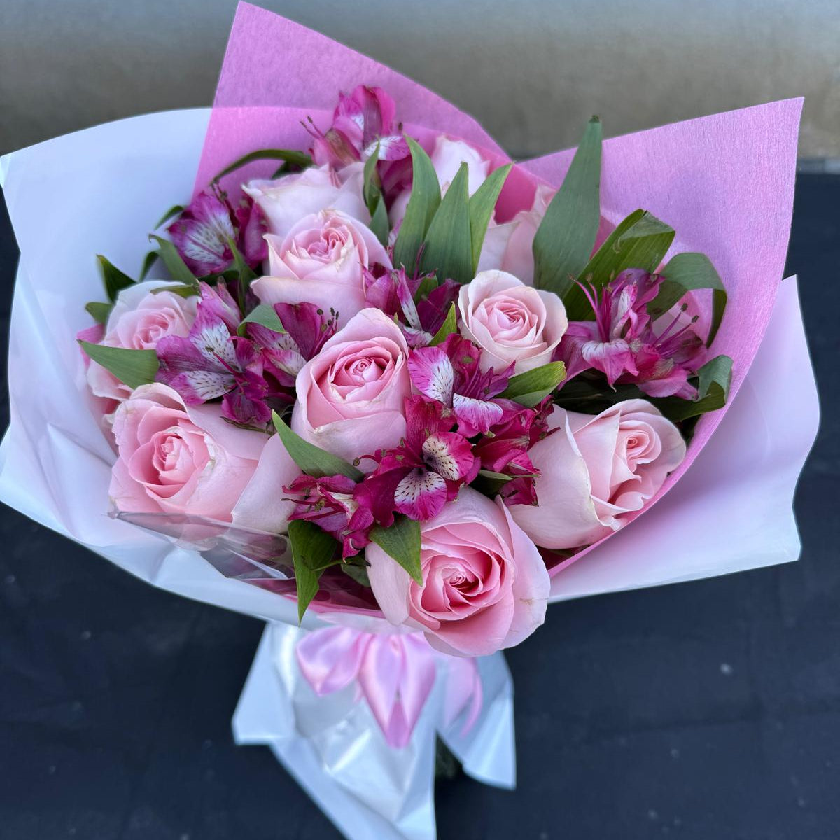 Blushing Grace Bouquet in one size only featuring blush pink roses and magenta alstroemeria wrapped in pink and white paper with satin ribbon by Nai Pascoal Bouquets